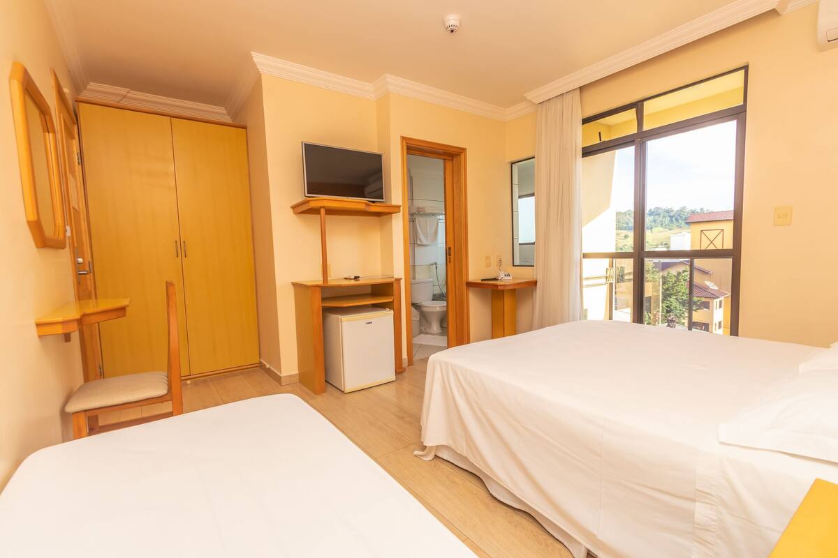 Ein Hotelzimmer mit zwei Betten, einem Fernseher, einer kleinen Kühlbox, einem Schreibtisch, einem Sessel und einem Balkon mit Blick.