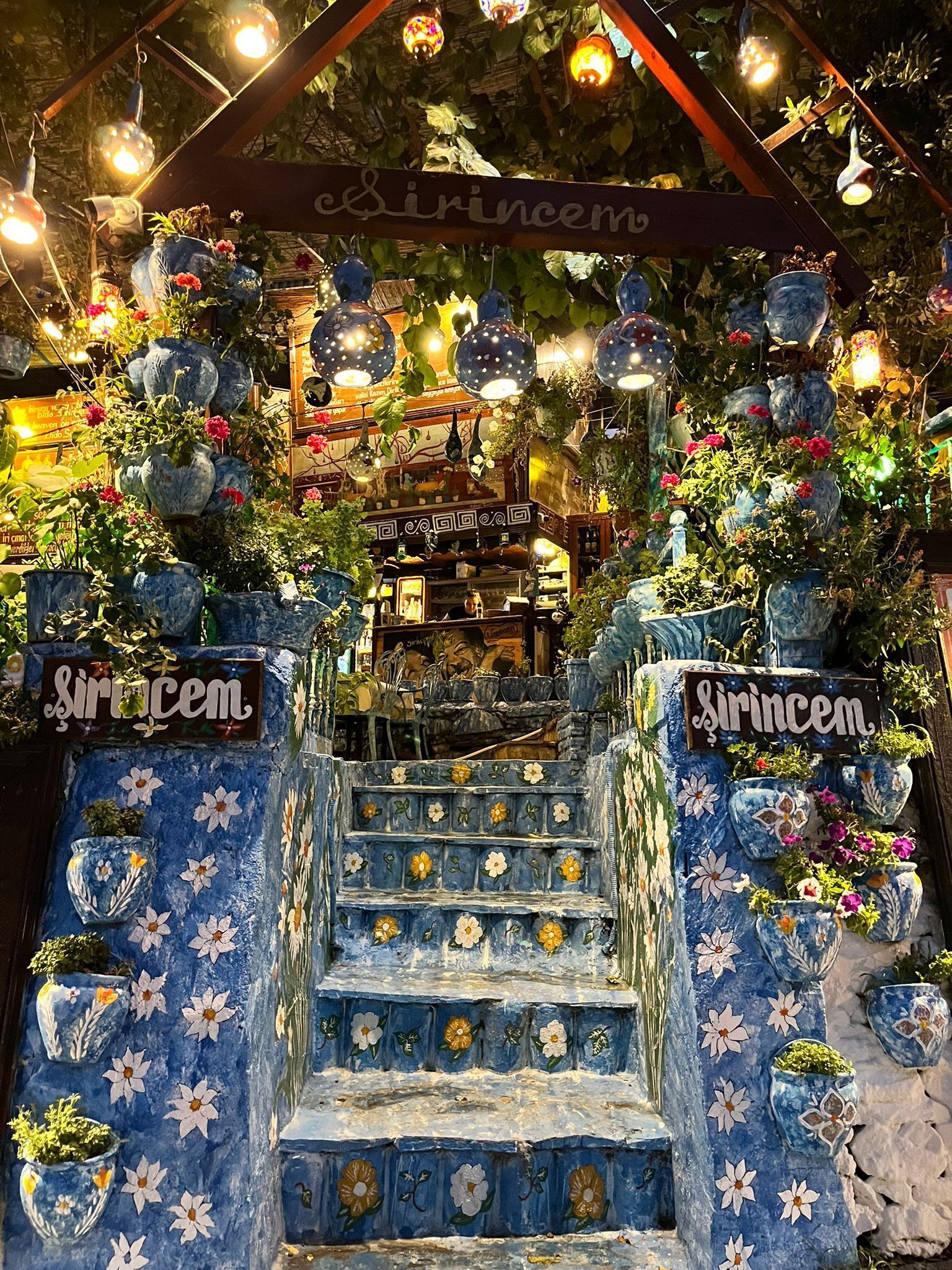 A staircase with floral patterns and hanging lights, leading to a shop area with a sign reading "Sirincem".