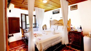 A bedroom with a wooden ceiling, a bed with white bedding and swan-shaped pillows, a wooden headboard, a nightstand with toiletries, and a window with curtains. - Sirincem Pension (Selçuk)