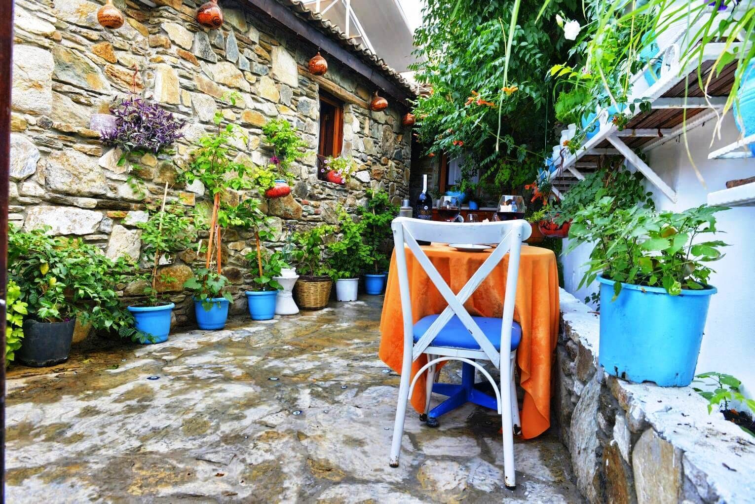 Un edificio de piedra con un patio estrecho, plantas en macetas y una mesa con una silla cubierta con un paño naranja.
