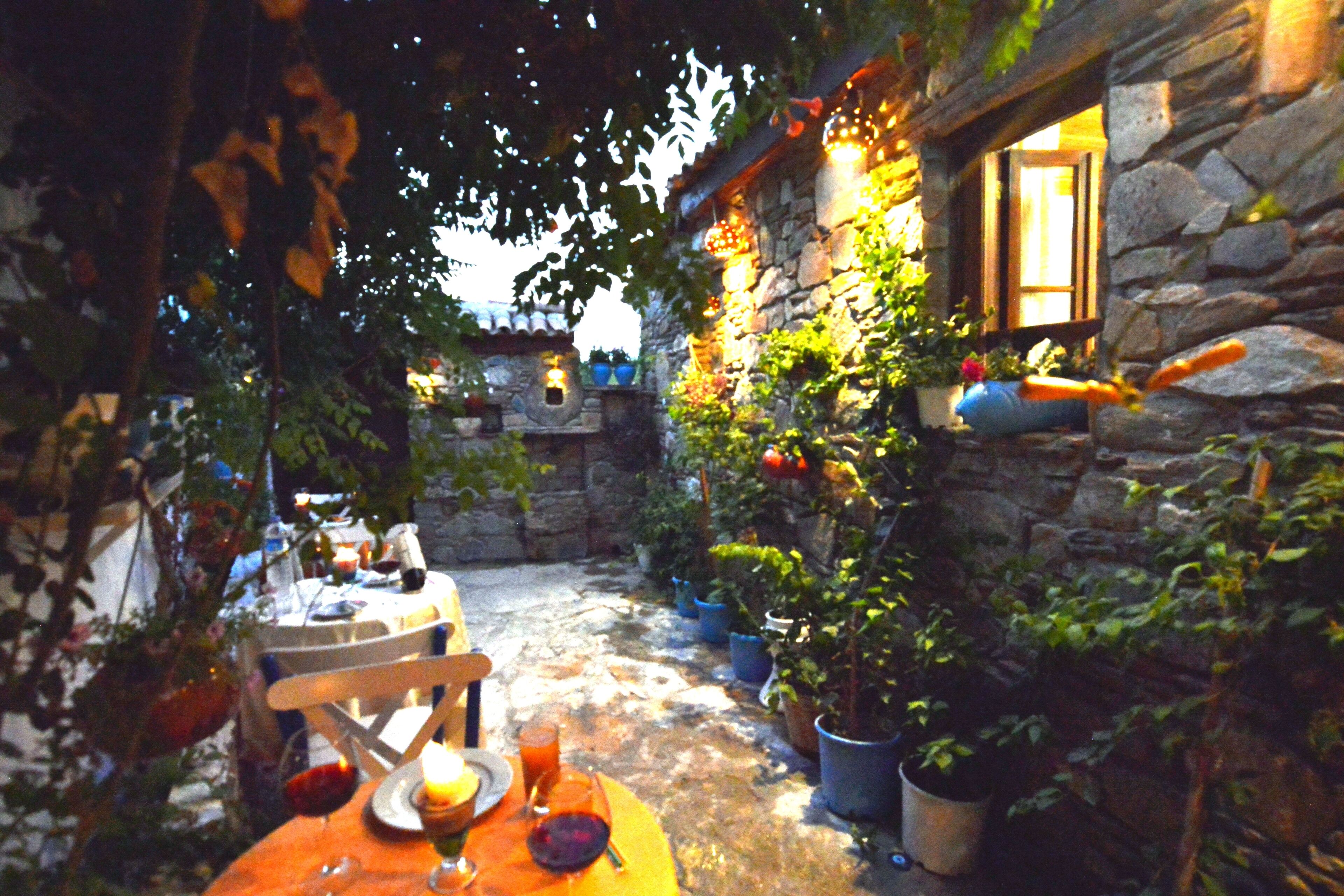 A stone building with a rustic outdoor seating area, potted plants, and a small table with a chair.