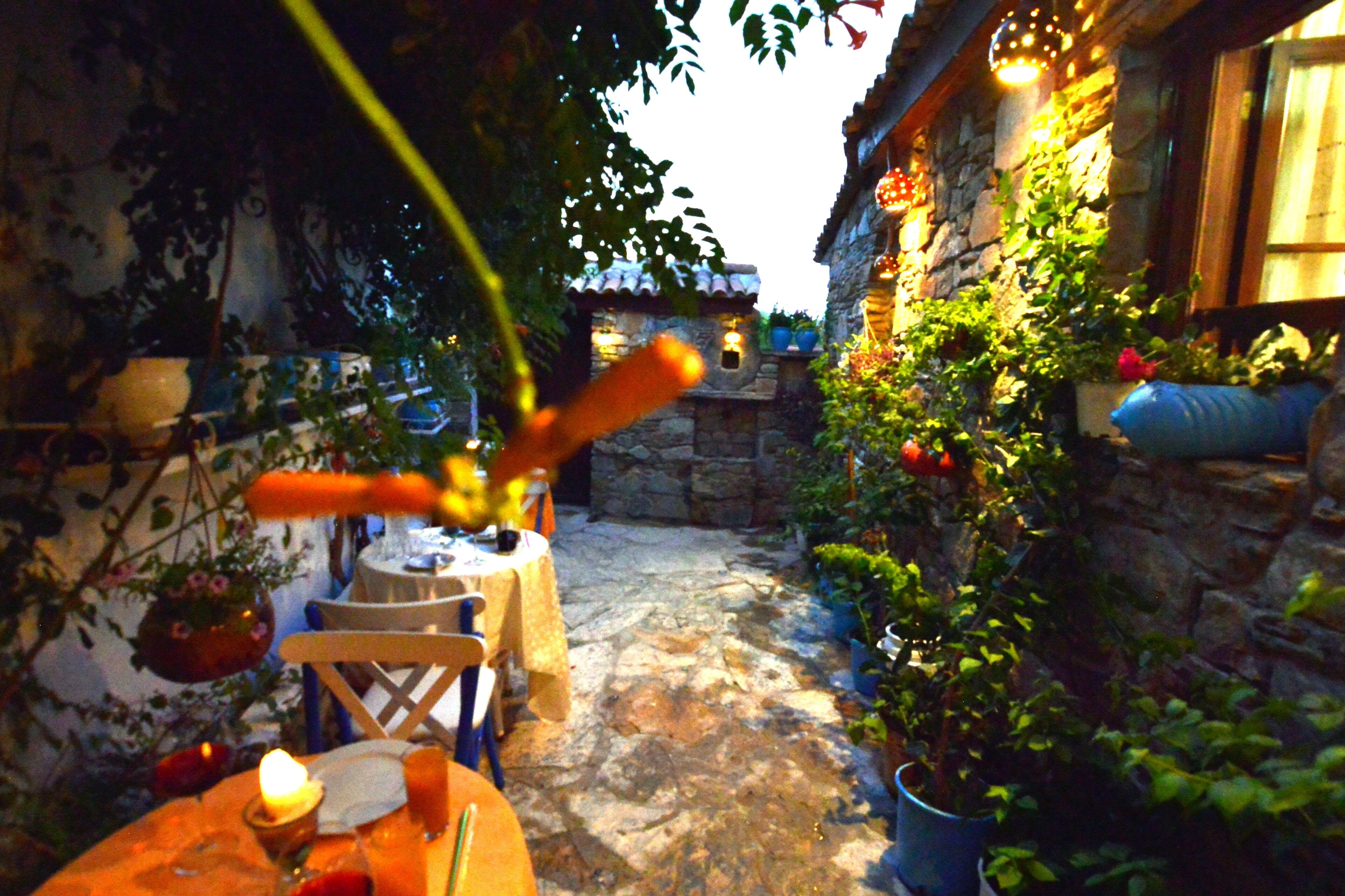 Un bâtiment en pierre à l’aspect rustique, un petit espace extérieur aménagé avec une table et des chaises, et des plantes en pot.