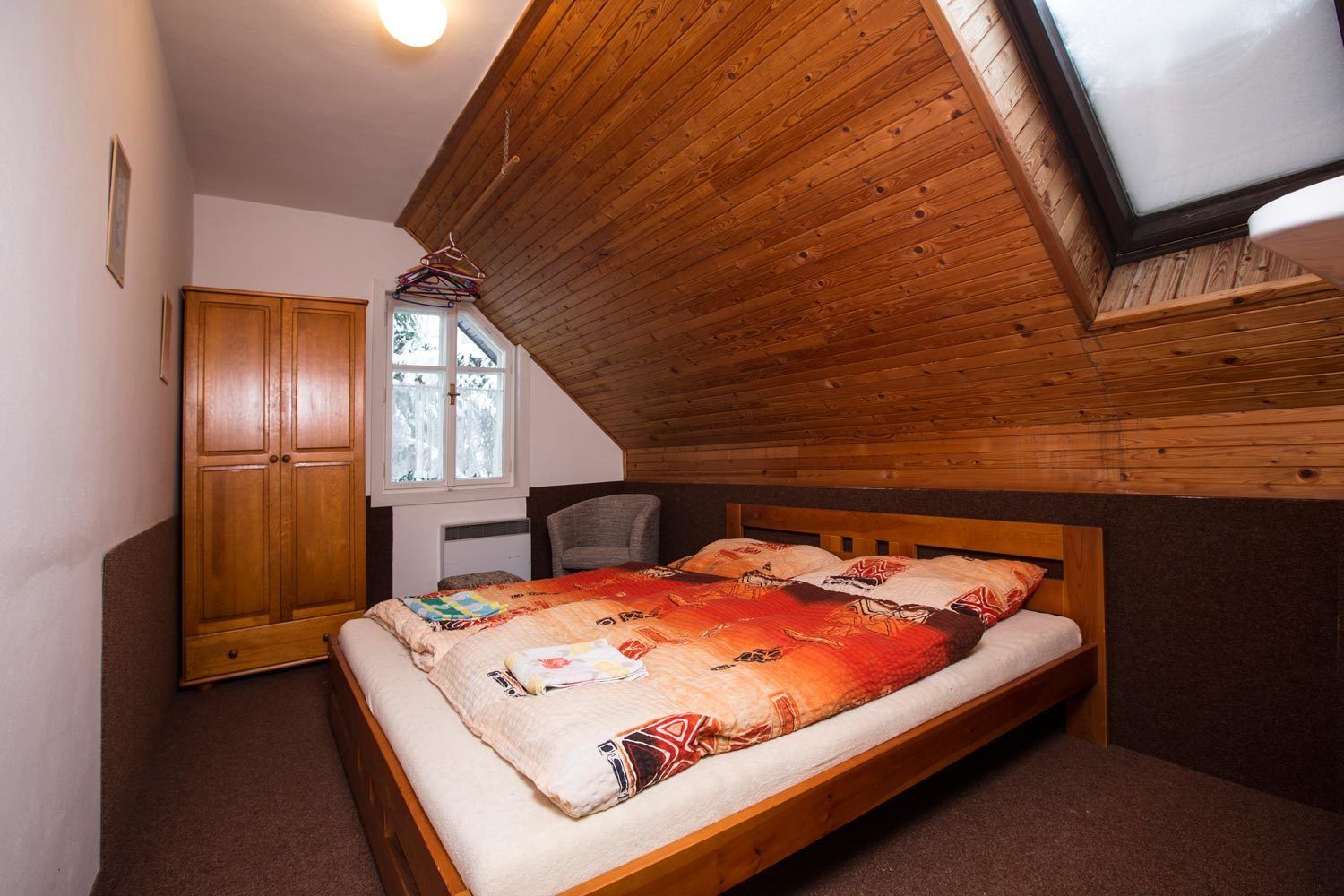 A bedroom with a sloped ceiling, a bed with a patterned blanket, a wooden wardrobe, and a window with a view of trees.