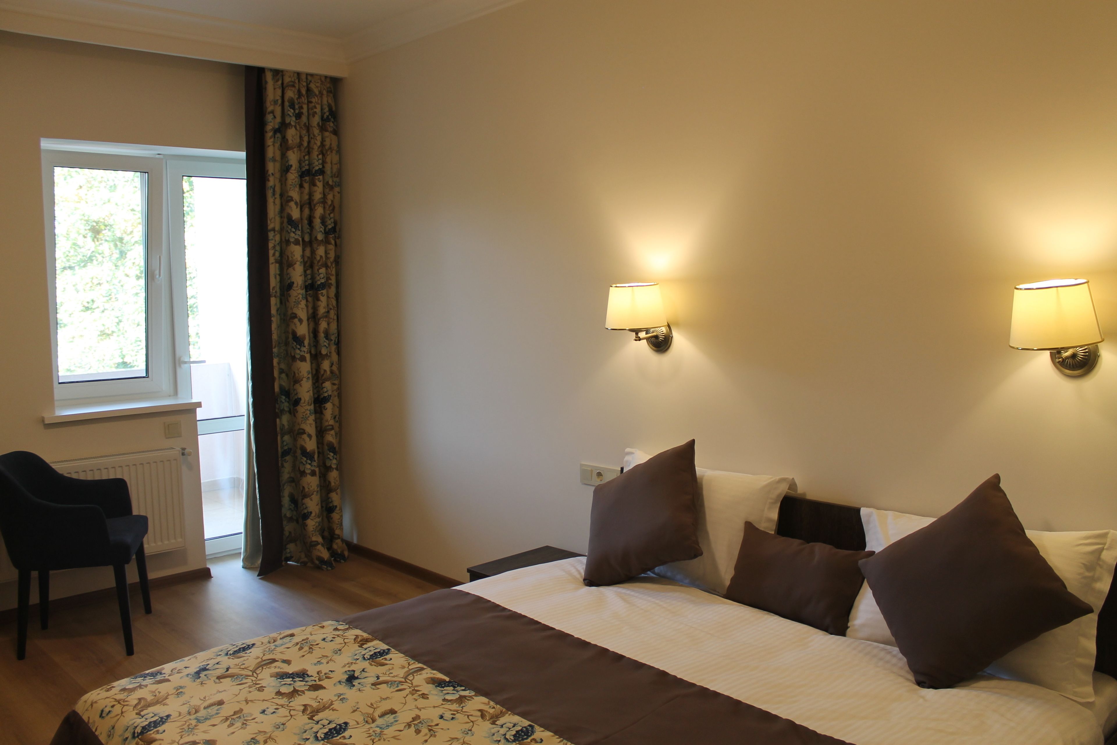 A hotel room with a bed, two wall-mounted lamps, a chair, a window with curtains, and a radiator.