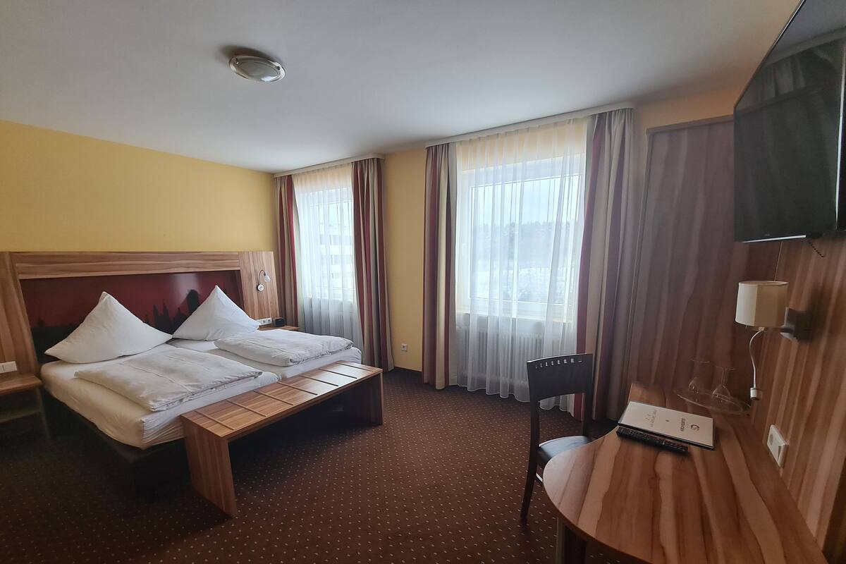 Ein Hotelzimmer mit Doppelbett, Schreibtisch mit Stuhl, Fernseher und einem Fenster mit Vorhängen.