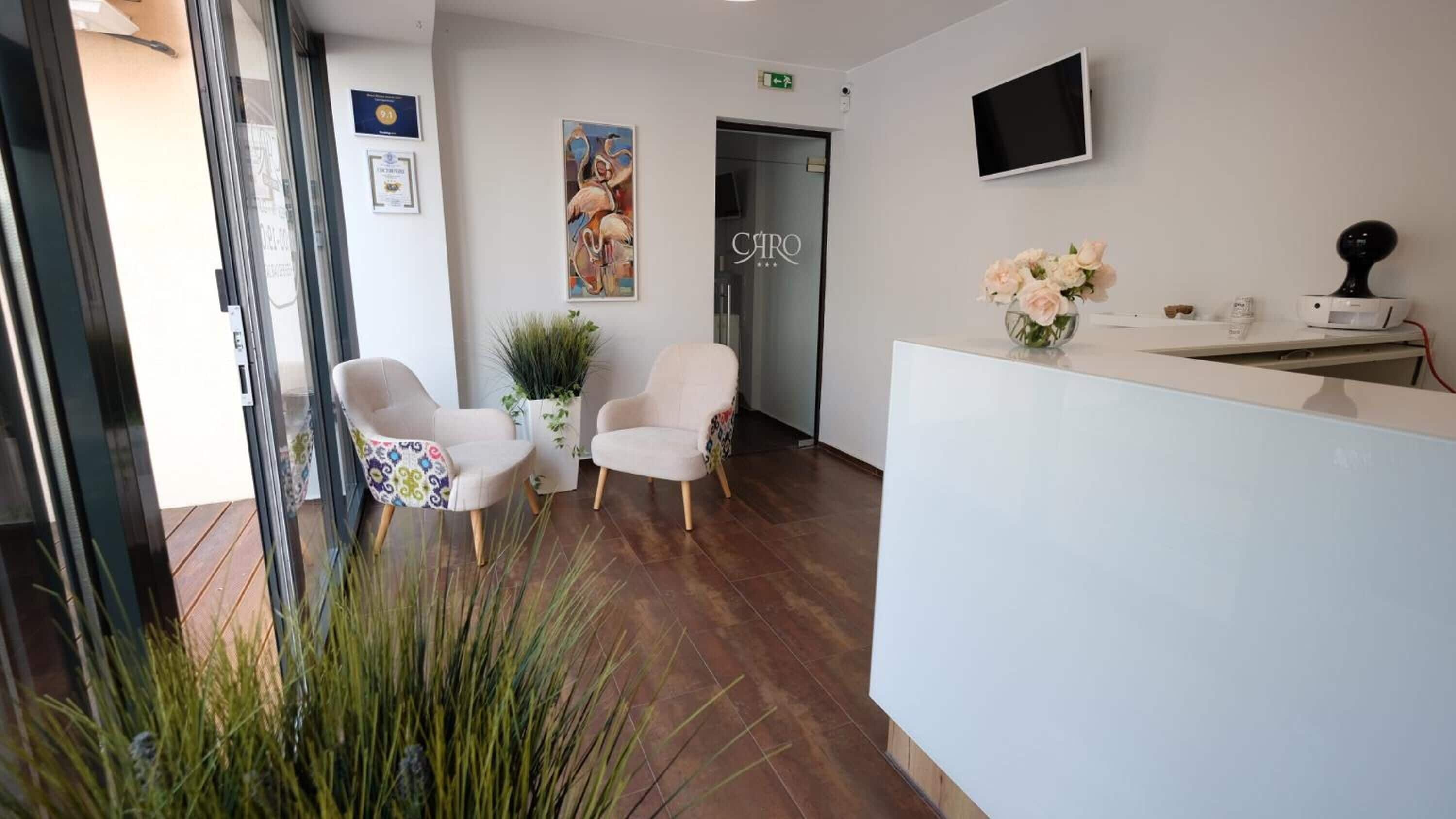 Una zona de recepción con un mostrador blanco, dos sillas blancas, una planta en maceta y un cuadro en la pared.