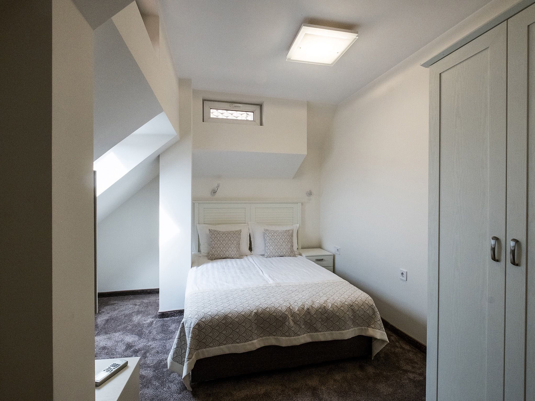 Un dormitorio con cama, mesita de noche, armario, tragaluz y suelo alfombrado.