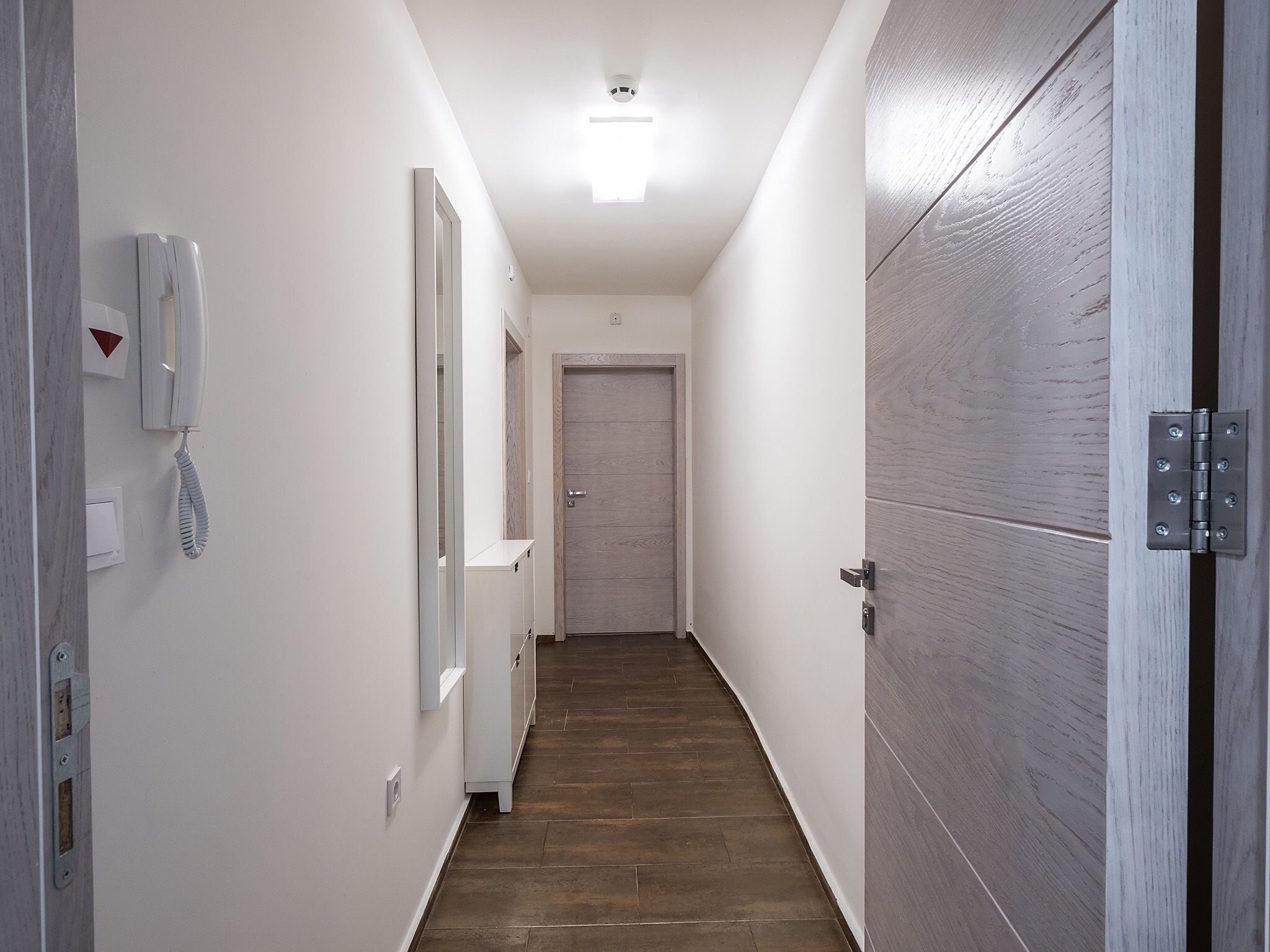 Un pasillo estrecho con piso de madera, una pared blanca con un teléfono y una puerta al final.