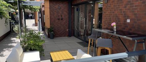 A modern outdoor seating area with a glass railing, a brick wall, and a door with a sign.