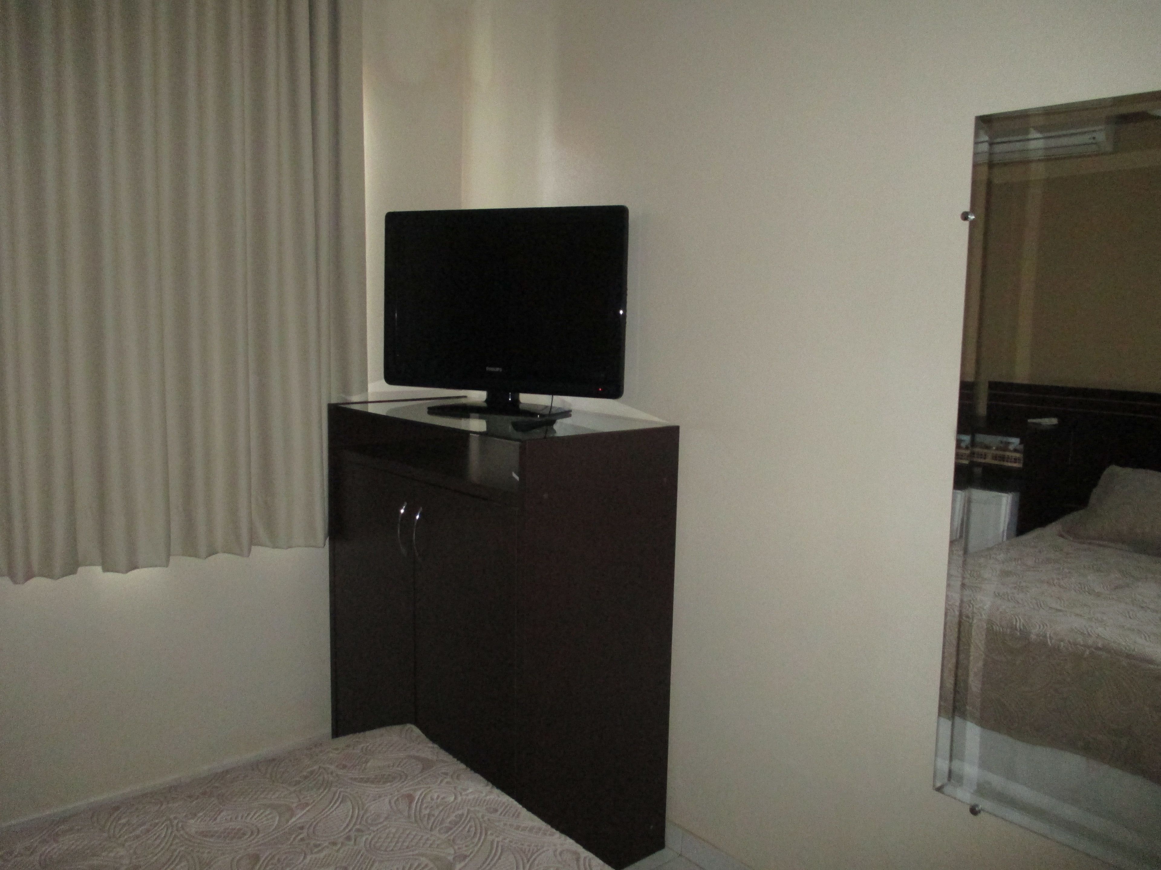 Ein Hotelzimmer mit einem dunklen Holzschrank, einem Fernseher darauf, einer gemusterten Bettdecke und einem Fenster mit Vorhängen.