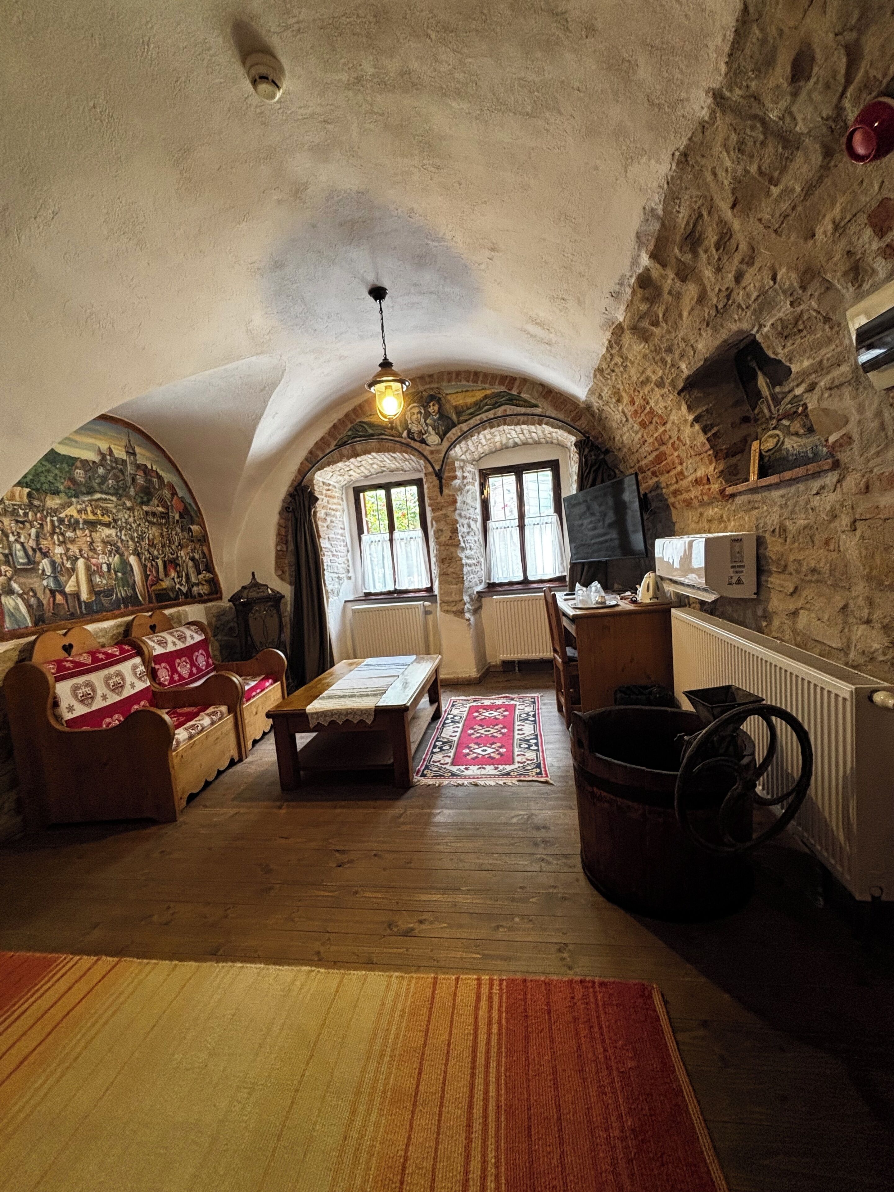 A cozy room with a wooden bench, a table, and a radiator. There is a mural on the wall and a window with curtains.