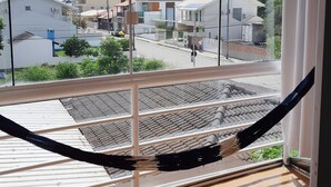 A hammock hanging by a window overlooking a street with buildings and greenery. - Canto do Pescador (Bombinhas)