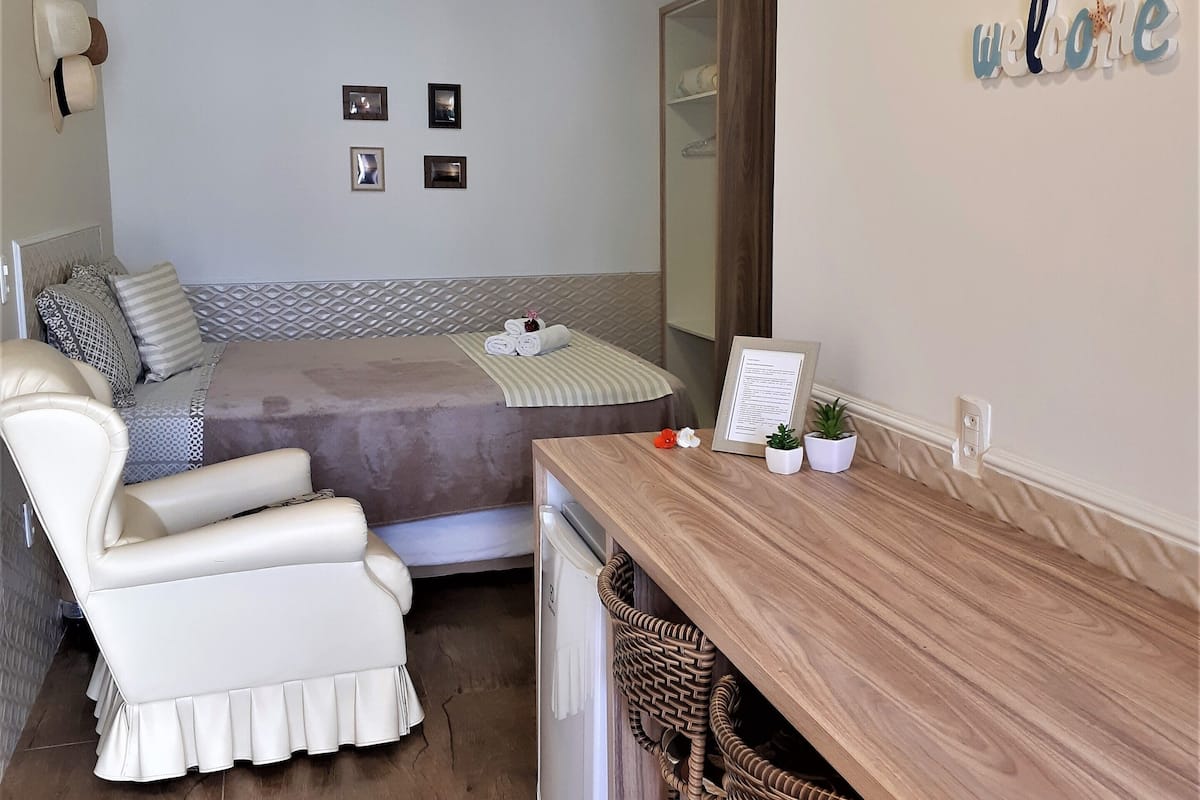 A cozy room with a bed, a white armchair, a wooden desk, and a wicker chair. There are two wall-mounted lights and a 'welcome' sign.