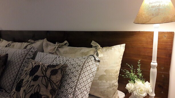 A neatly made bed with patterned pillows, a white vase with flowers, and a lamp on a wooden nightstand. - Canto do Pescador (Bombinhas)