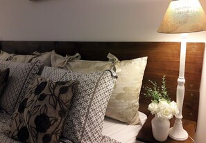 Uma cama bem arrumada com almofadas estampadas, um vaso branco com flores e um abajur em uma mesa de cabeceira de madeira.