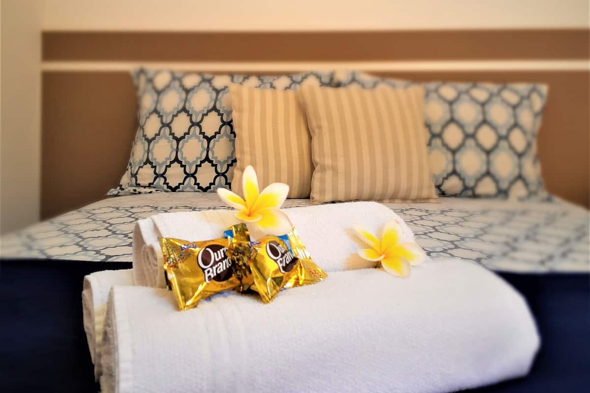 A neatly made bed with a patterned bedspread, two pillows, a folded white towel, and two wrapped chocolate bars on top.