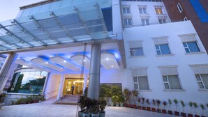 Una entrada de hotel moderna con un toldo de cristal, una escalera que lleva al vestíbulo y una fila de plantas en macetas a lo largo del acceso.