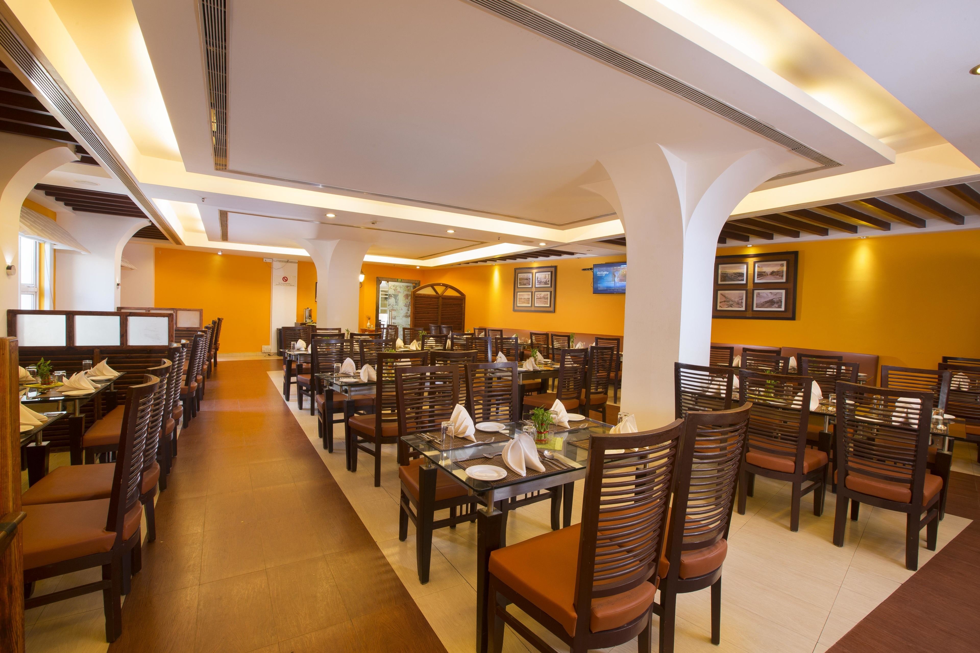 Un restaurante espacioso con varias mesas y sillas, techos altos con iluminación empotrada y una paleta de colores cálida.