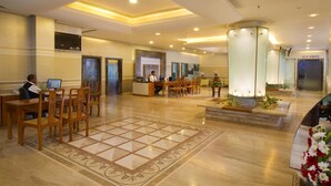 Un vestíbulo de hotel con mostrador de recepción, varias sillas de madera, suelo de baldosas y un pilar de cristal con luces.
