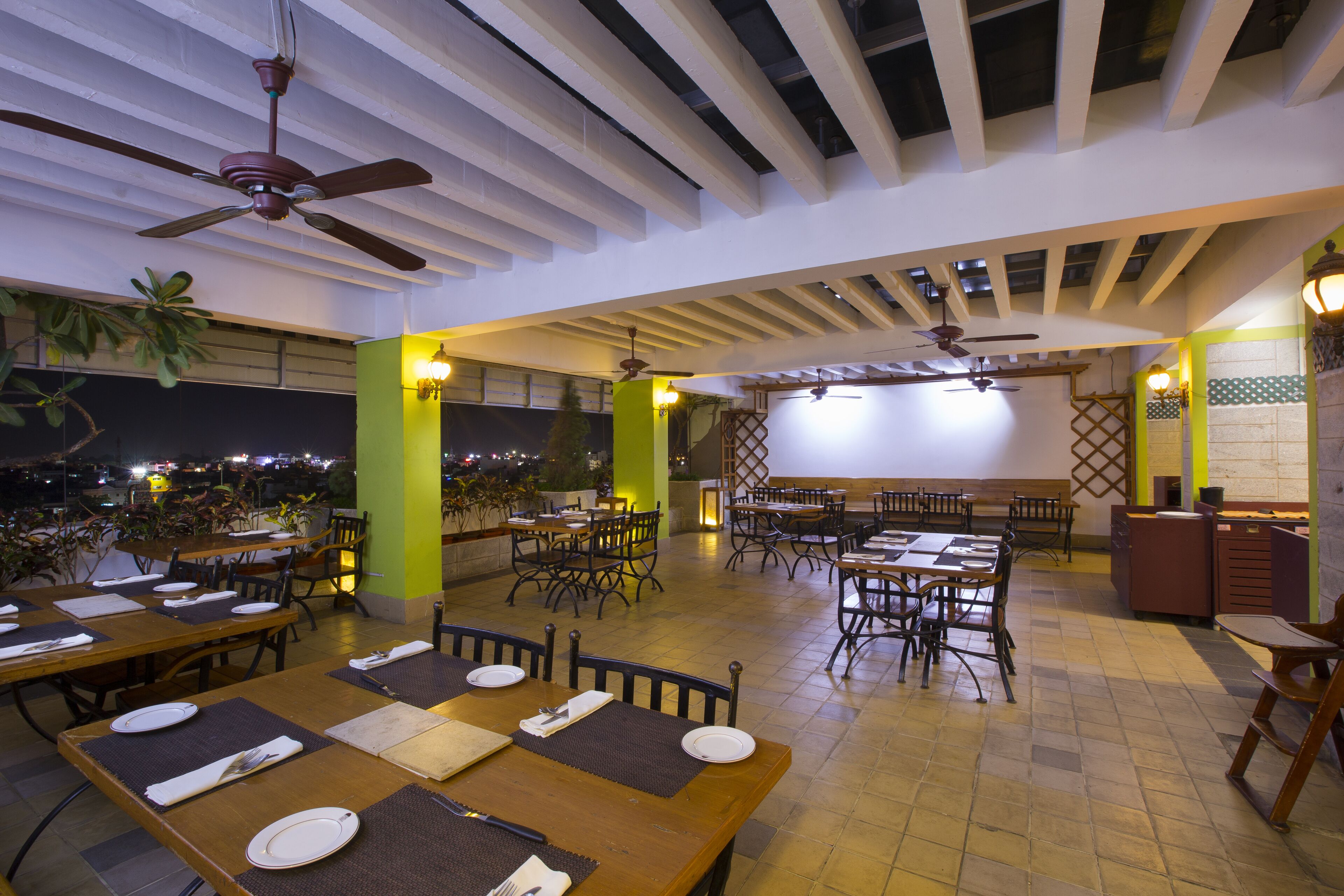 An outdoor restaurant with multiple tables and chairs, ceiling fans, and a view of the city at night.