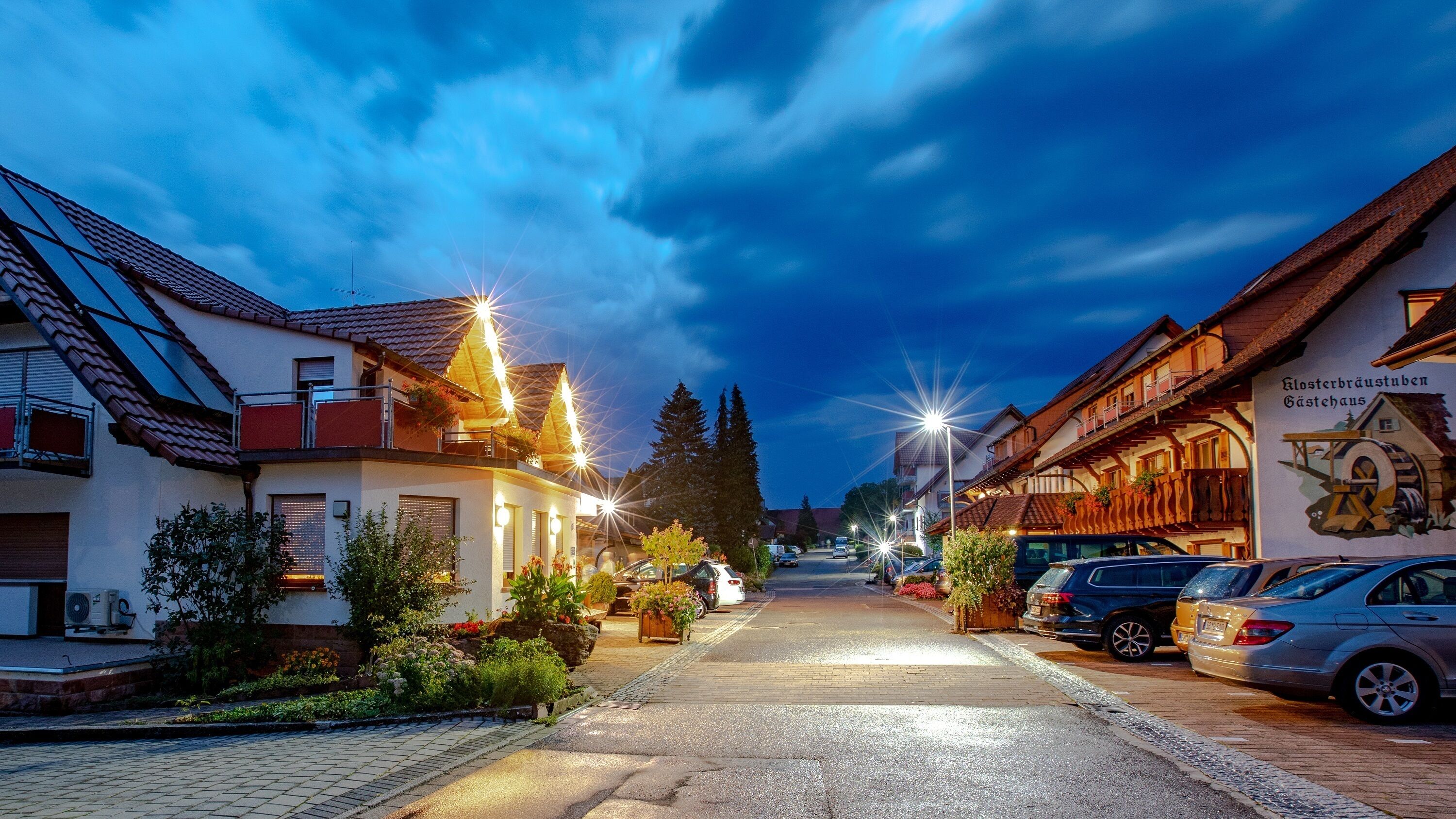 Eine Straße gesäumt von traditionellen Gebäuden, ein asphaltierter Weg und parkende Autos.