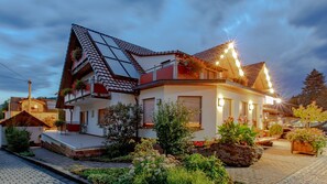 Una casa de dos pisos con techo inclinado, un balcón con barandillas rojas y un jardín con diversas plantas y flores.