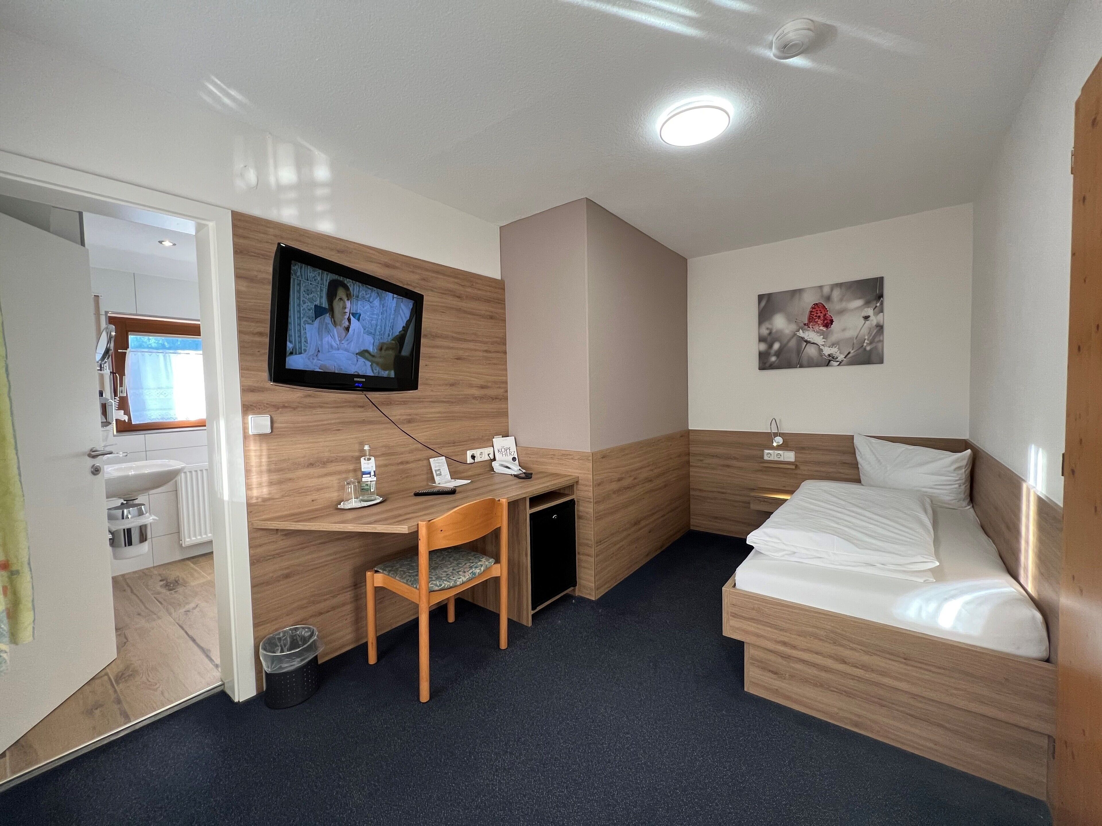 Une chambre d’hôtel de taille modeste, comprenant un lit, un bureau avec une chaise, une télévision, une petite salle de bain et un tableau au mur.