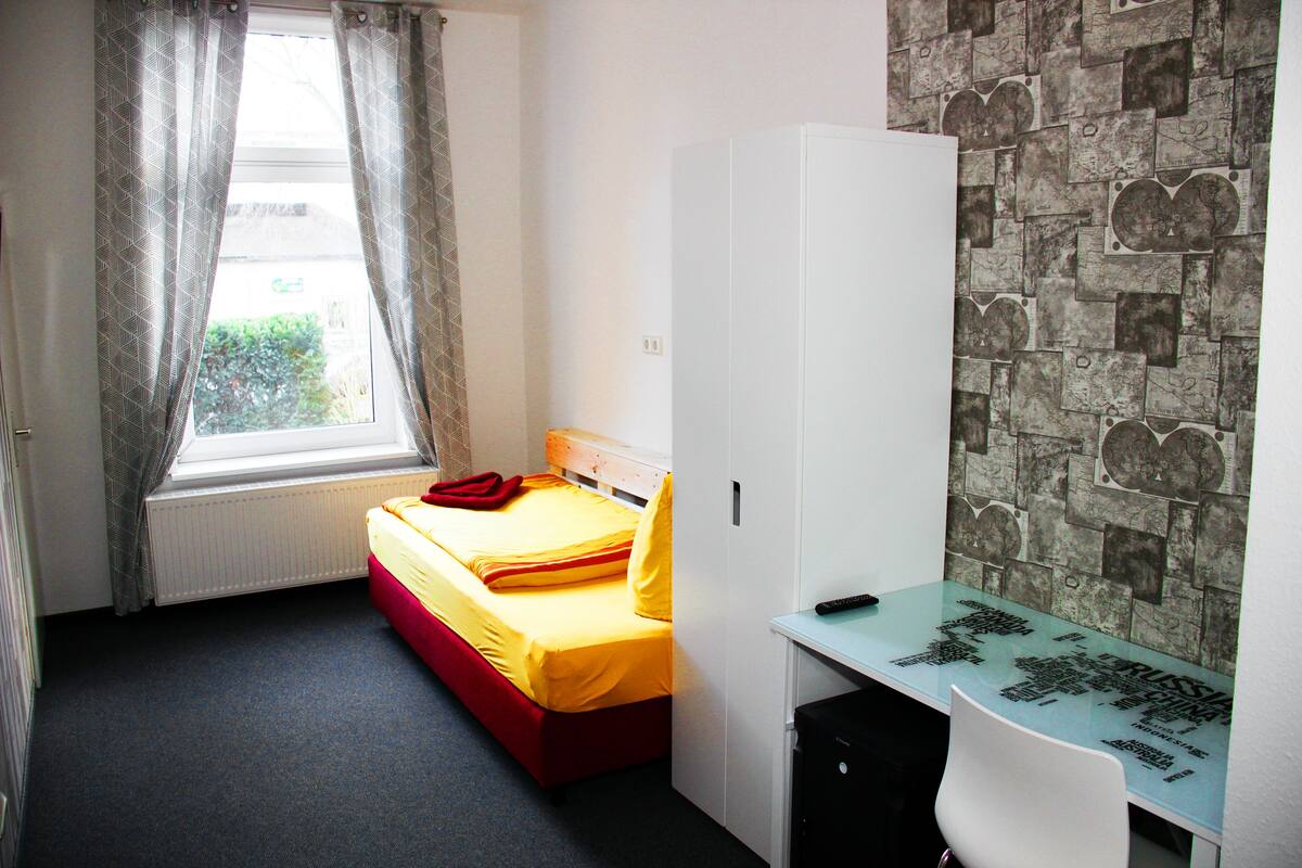 A bedroom with a bed, a wardrobe, a desk, and a window with curtains.