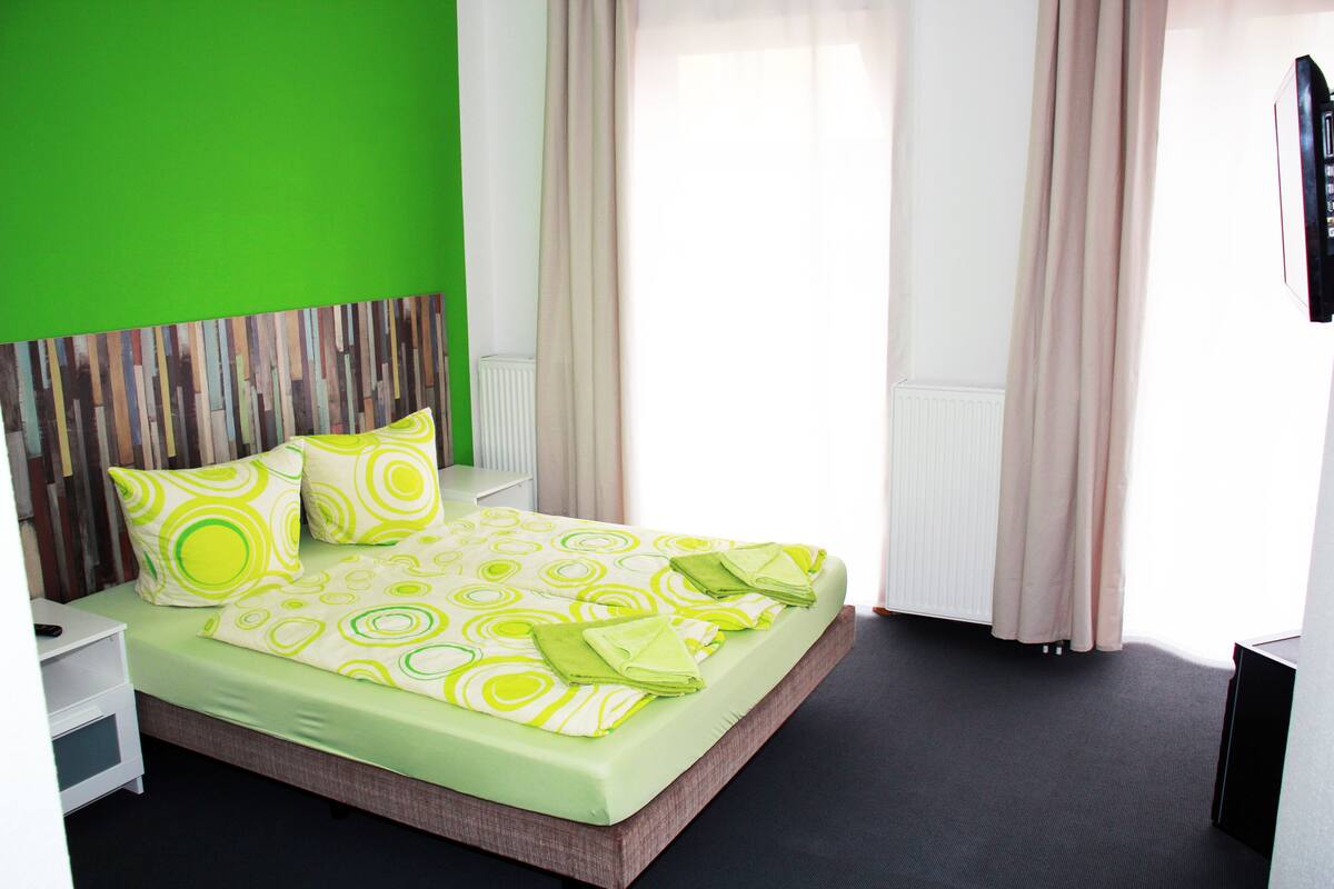 A modern hotel room with a green wall, a wooden headboard, a neatly made bed with green and white bedding, and a large window with curtains.