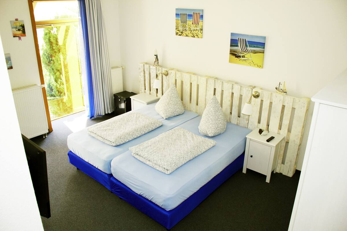 A double bed with a blue mattress and white pillows, a white bedside table with a lamp, and a small nightstand.