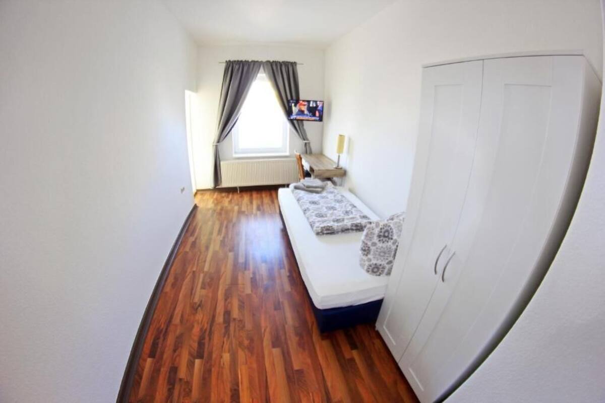 A narrow room with a wooden floor, a white wardrobe, a bed with patterned bedding, a television on a table, and a window with curtains.