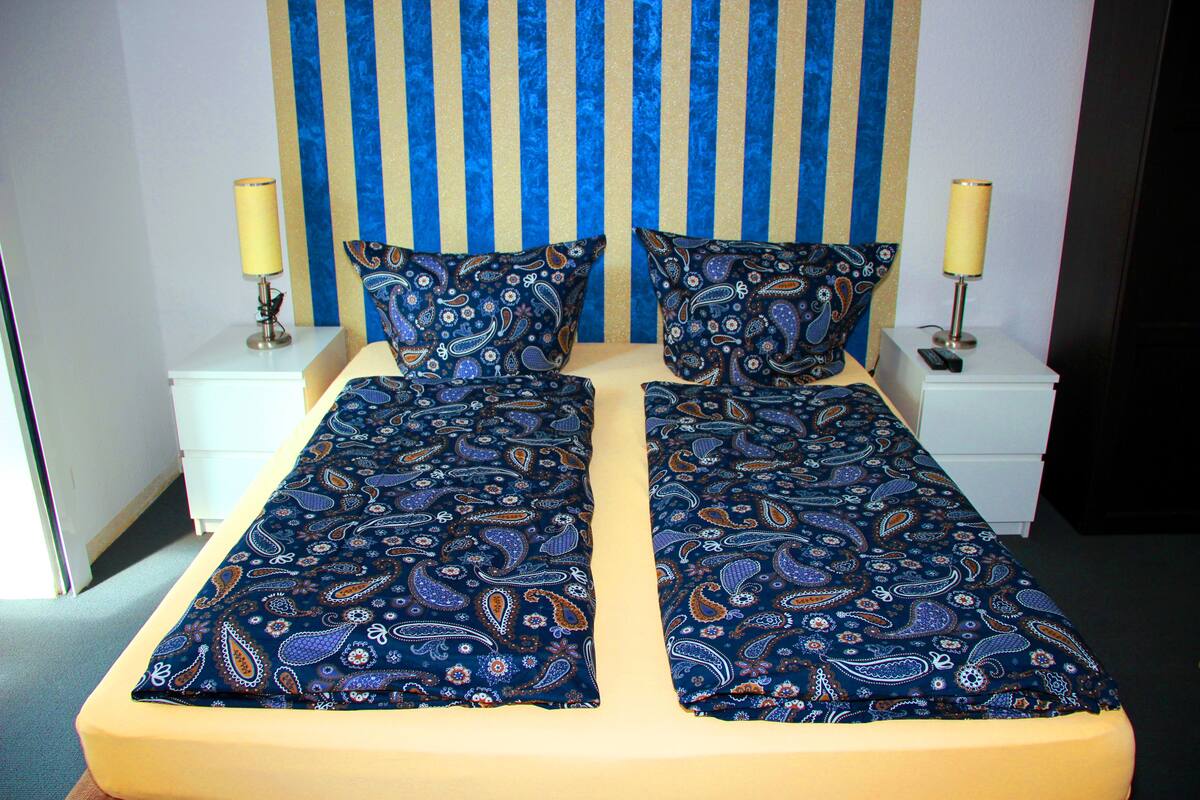 A double bed with a blue paisley bedspread, two bedside lamps, and a striped headboard.