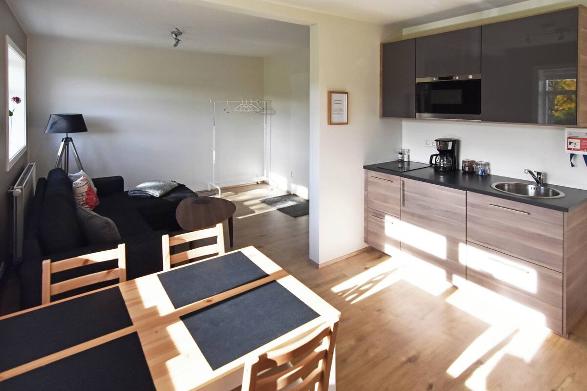 Een moderne keuken met houten kastjes, een magnetron en een koffiezetapparaat. Er is een eethoek met een tafel en stoelen, en een zithoek met een bank en een lamp.