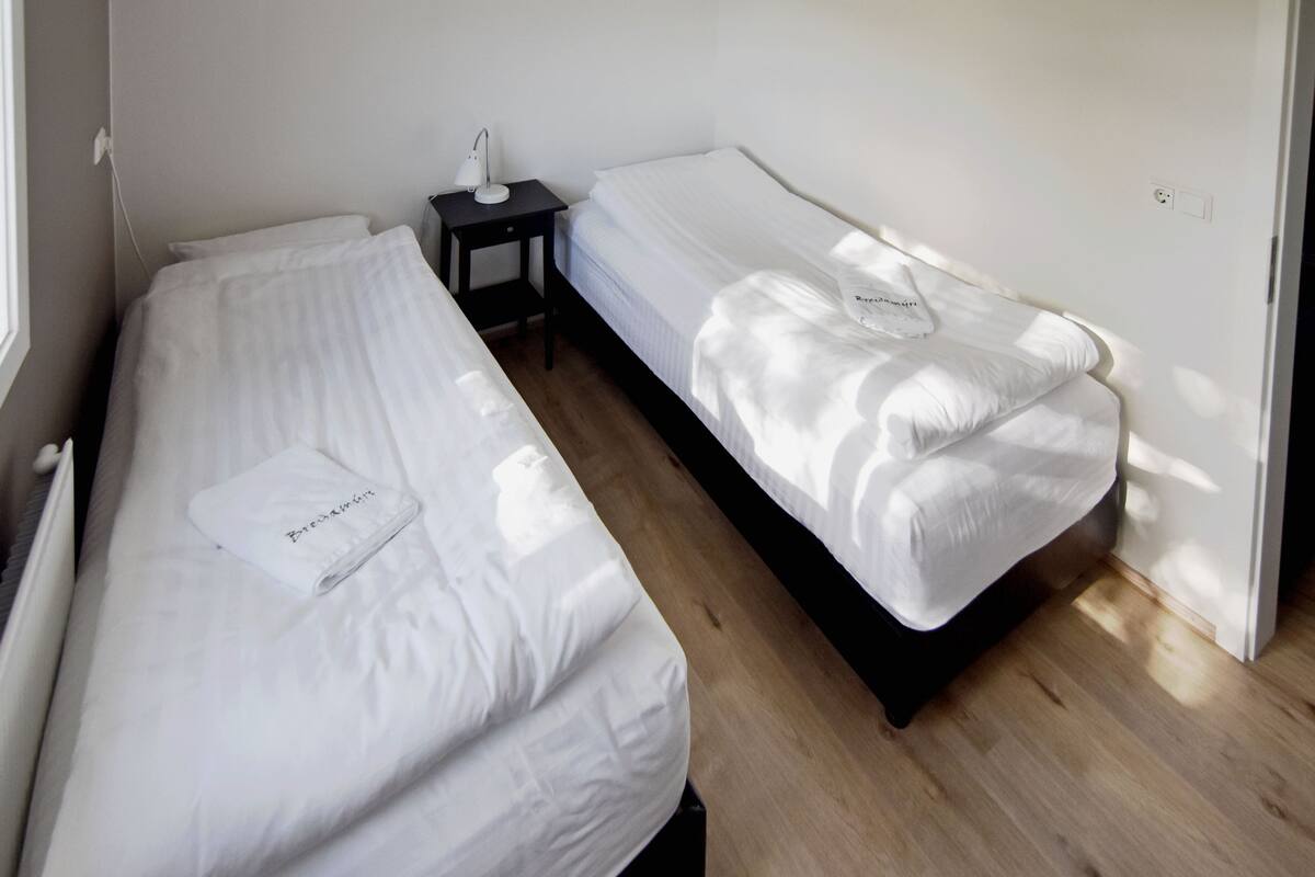 Een kamer met twee eenpersoonsbedden, een klein nachtkastje met een lamp, en een witte wand.
