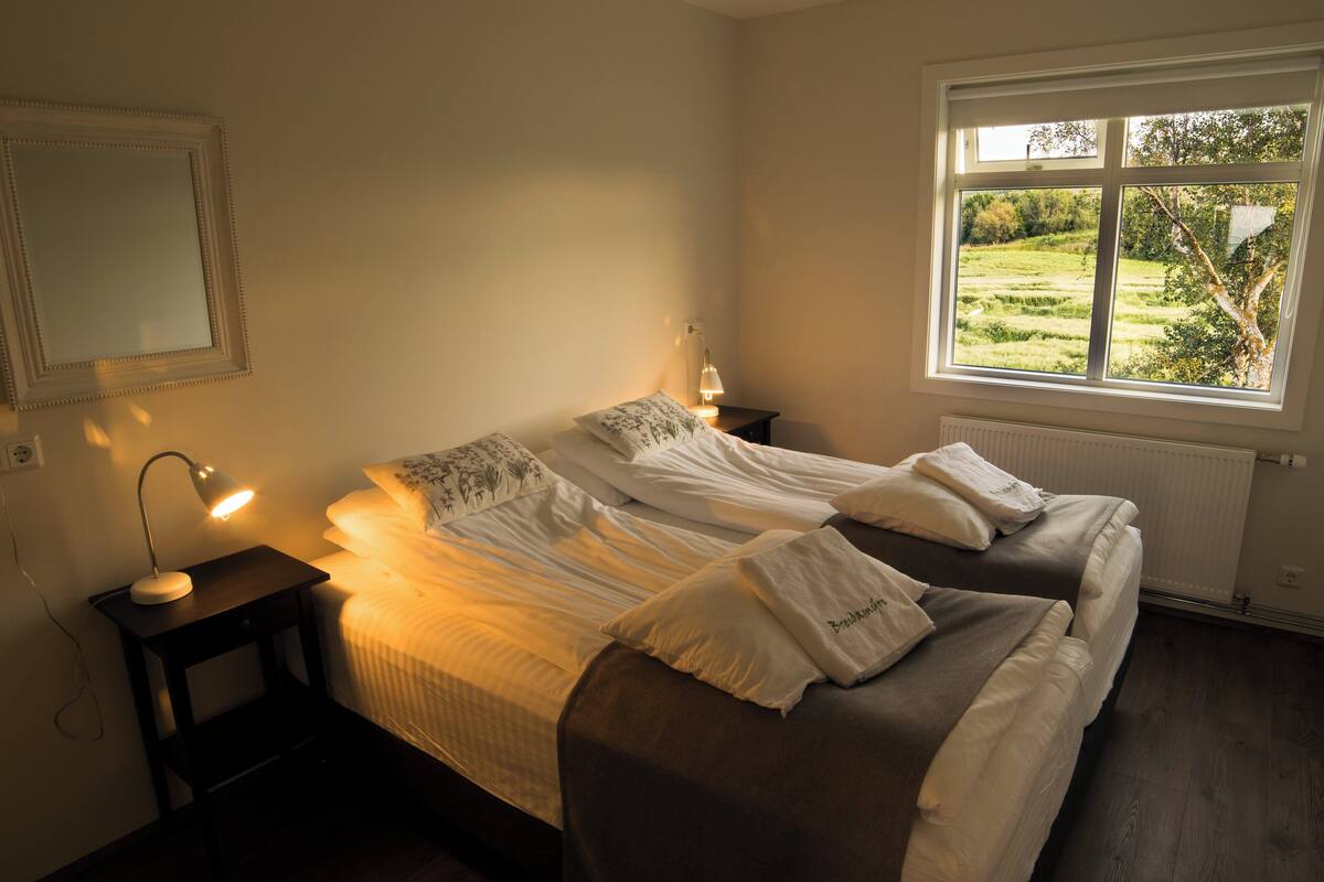 Een slaapkamer met een bed, nachtkastjes, een lamp, een raam met uitzicht op groen en een radiator.
