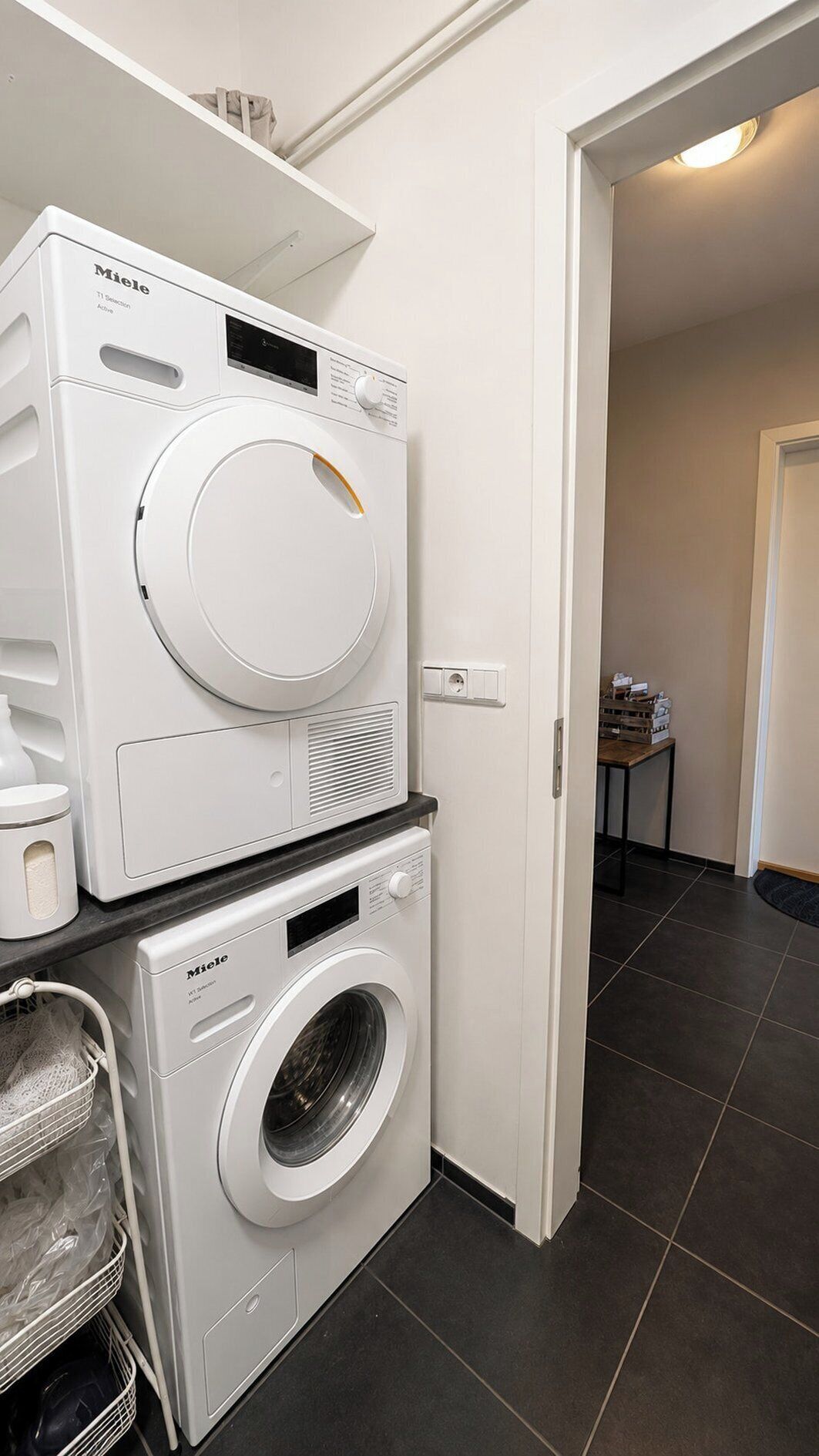 A laundry room with two white Miele washing machines, a small table with items, and a door leading to another room.