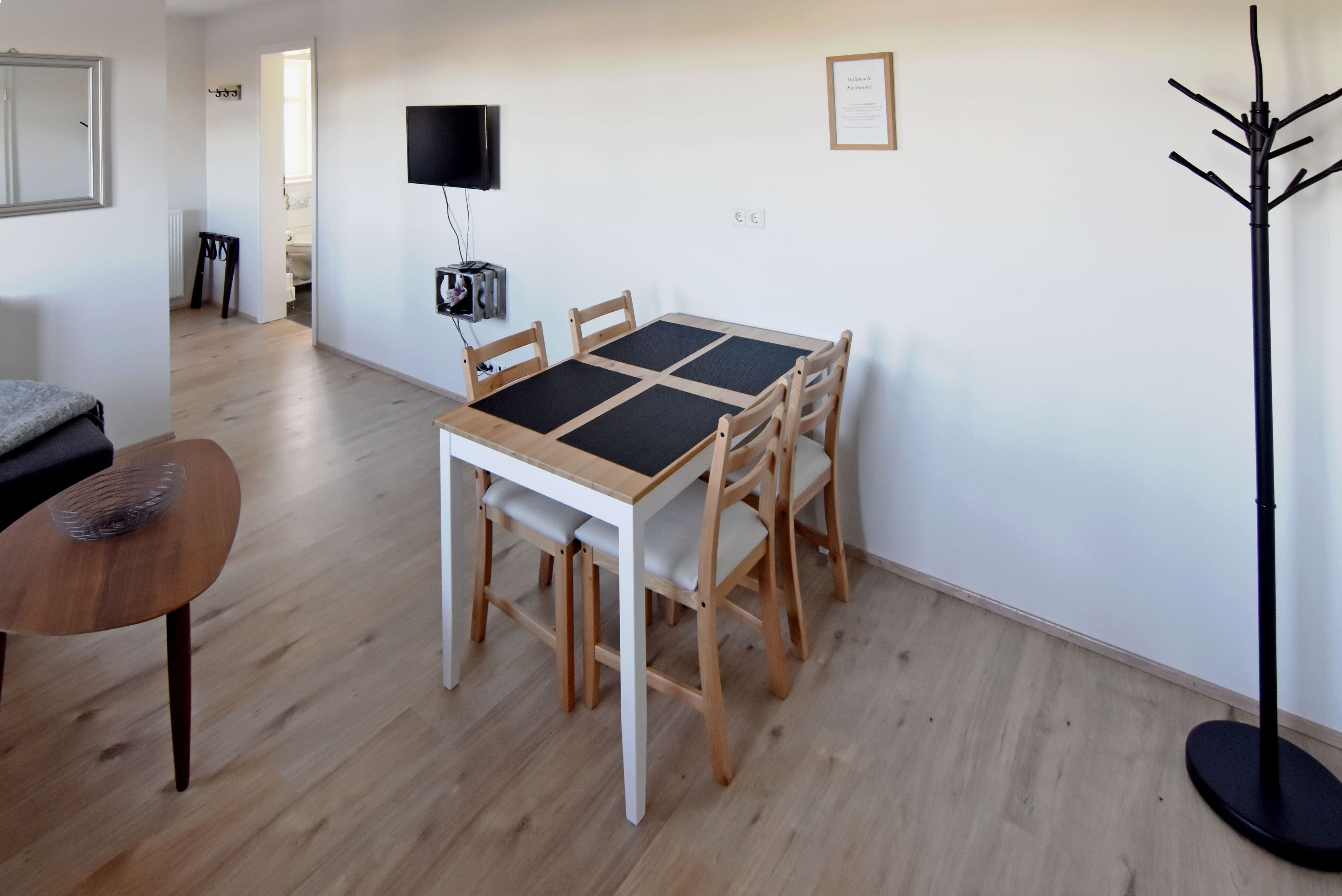 A dining area with a wooden table and chairs, a television mounted on the wall, and a coat rack.