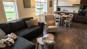 A modern living room with a dark grey sofa, a decorative chair, and a dining table with chairs. A large window offers a view of the outdoors. - Breidamyri Farm Apartments (Laugar)