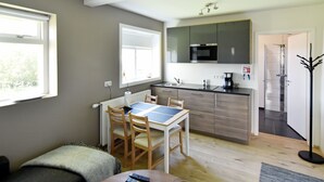 A compact living space with a dining table, chairs, a sofa, a kitchen area with a microwave, and a view of a garden through the window. - Breidamyri Farm Apartments (Laugar)