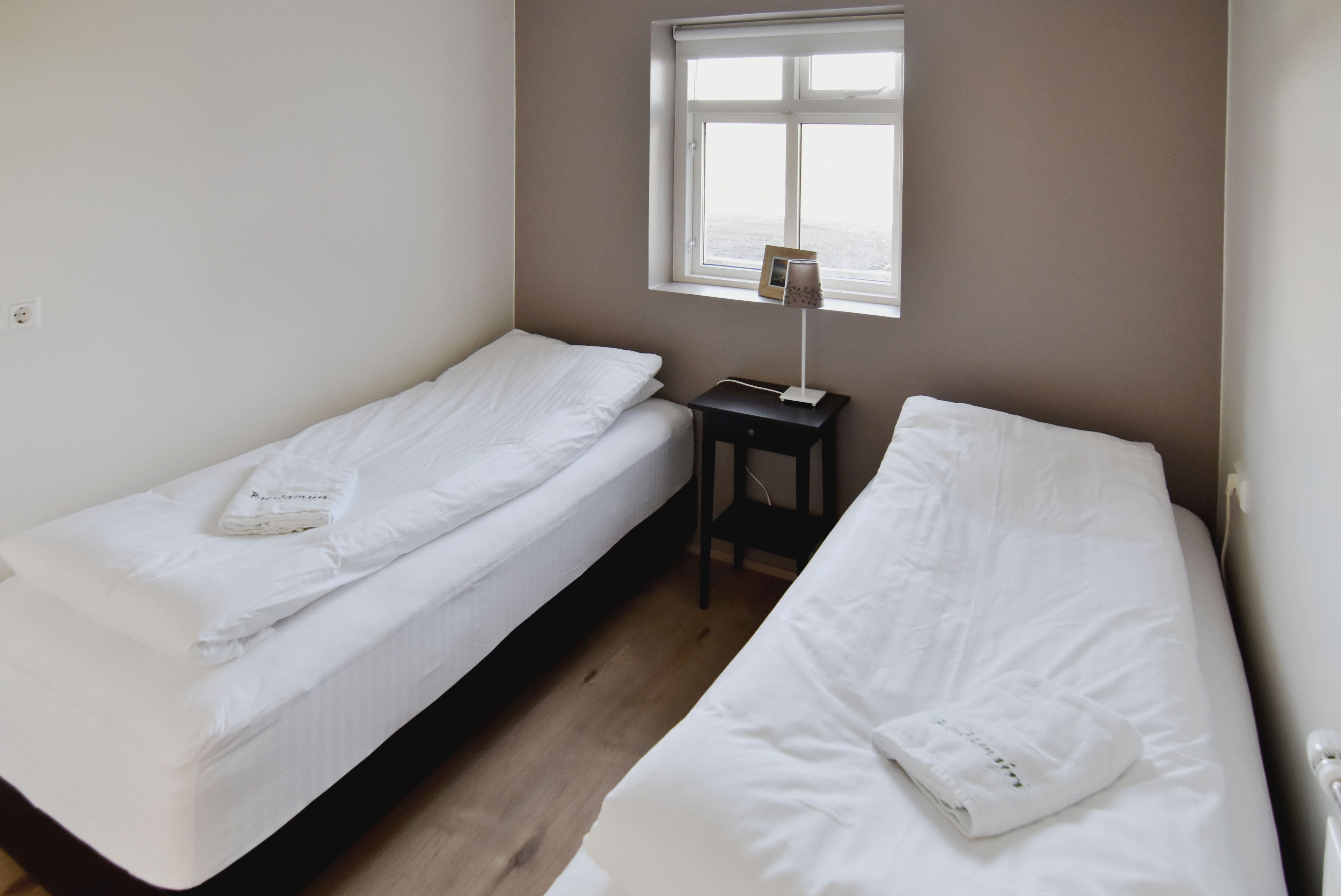 A small, clean room with two single beds, a nightstand with a lamp, and a window with a view.