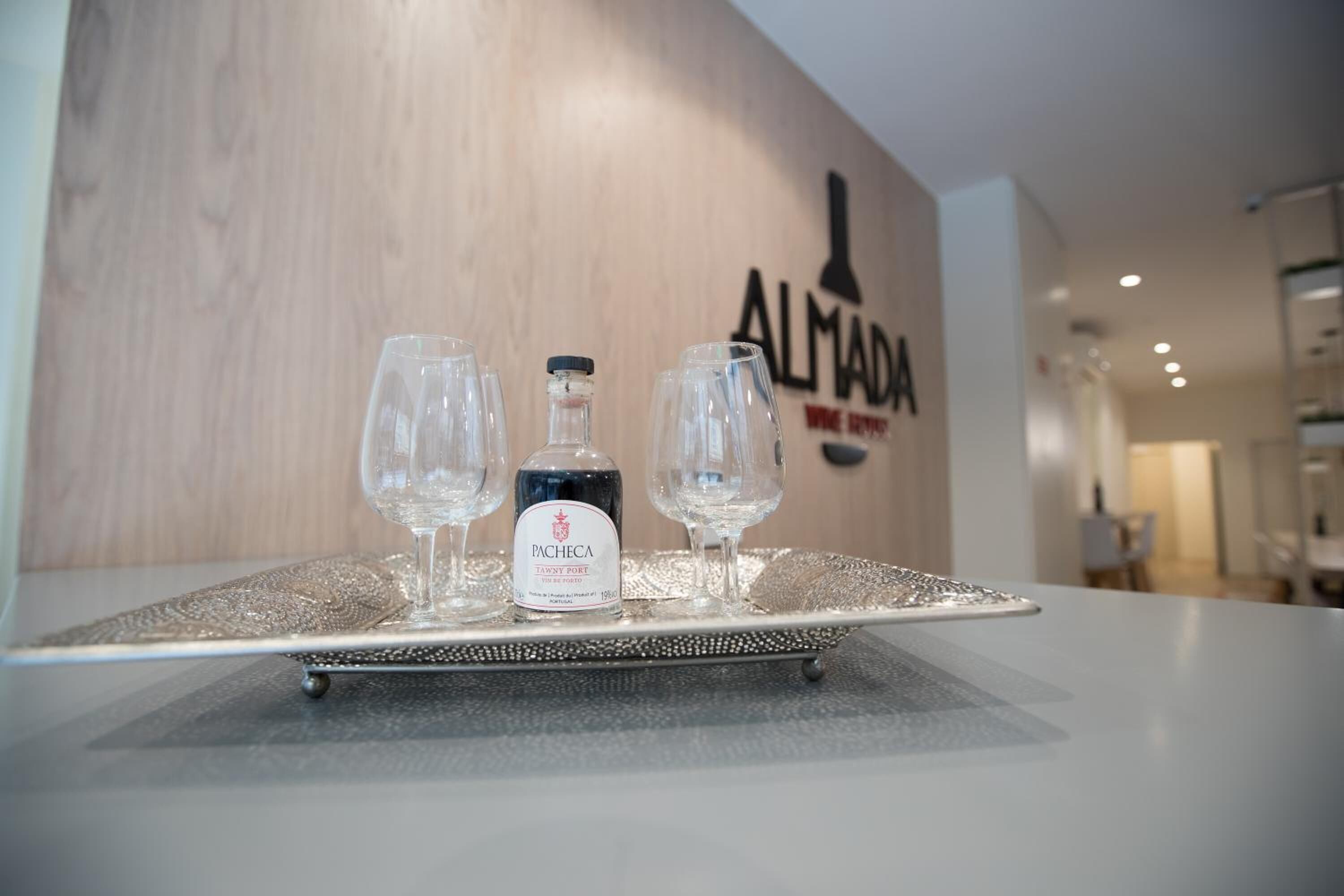 Un plateau avec une bouteille de porto tawny de la marque Pacheca et deux verres à vin vides est posé sur une table devant un mur orné du logo « ALMA DA WINE SHOP ».