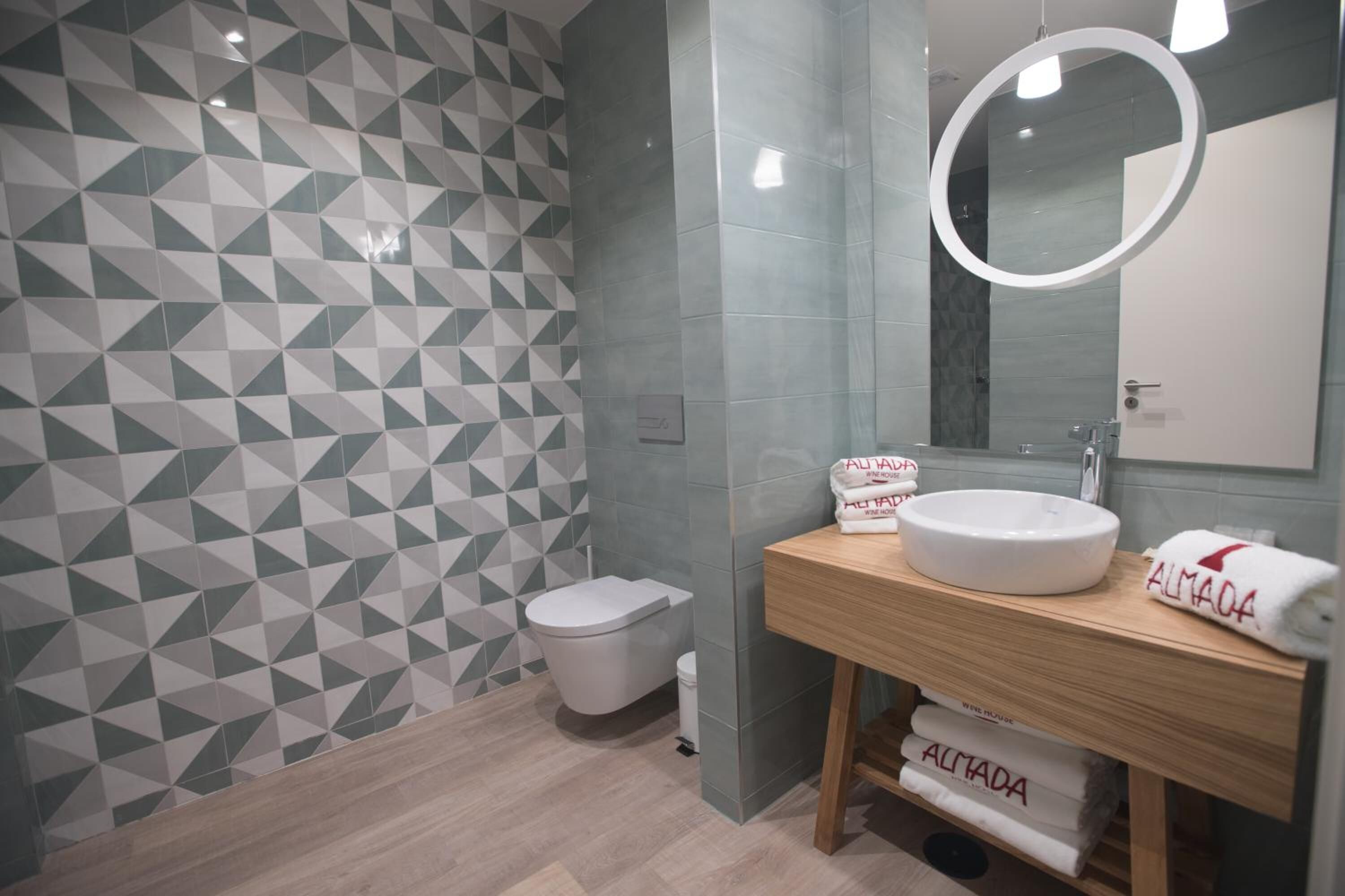 A modern bathroom with geometric patterned tiles, a wooden vanity, a round mirror, and a toilet.