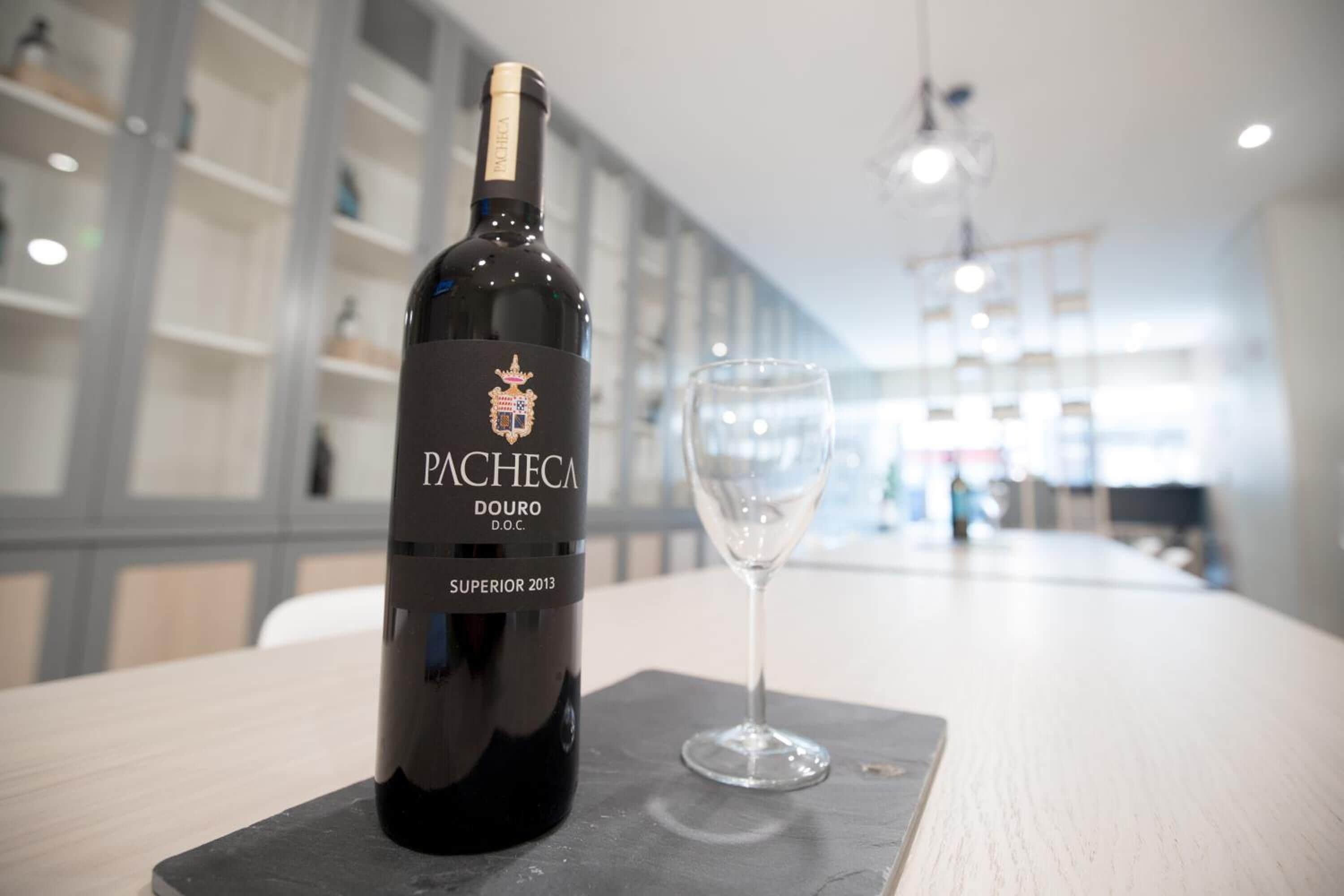 A bottle of Pacheca Douro D.O.C. Superior 2013 wine and a wine glass on a table in a modern, well-lit room with shelves in the background.
