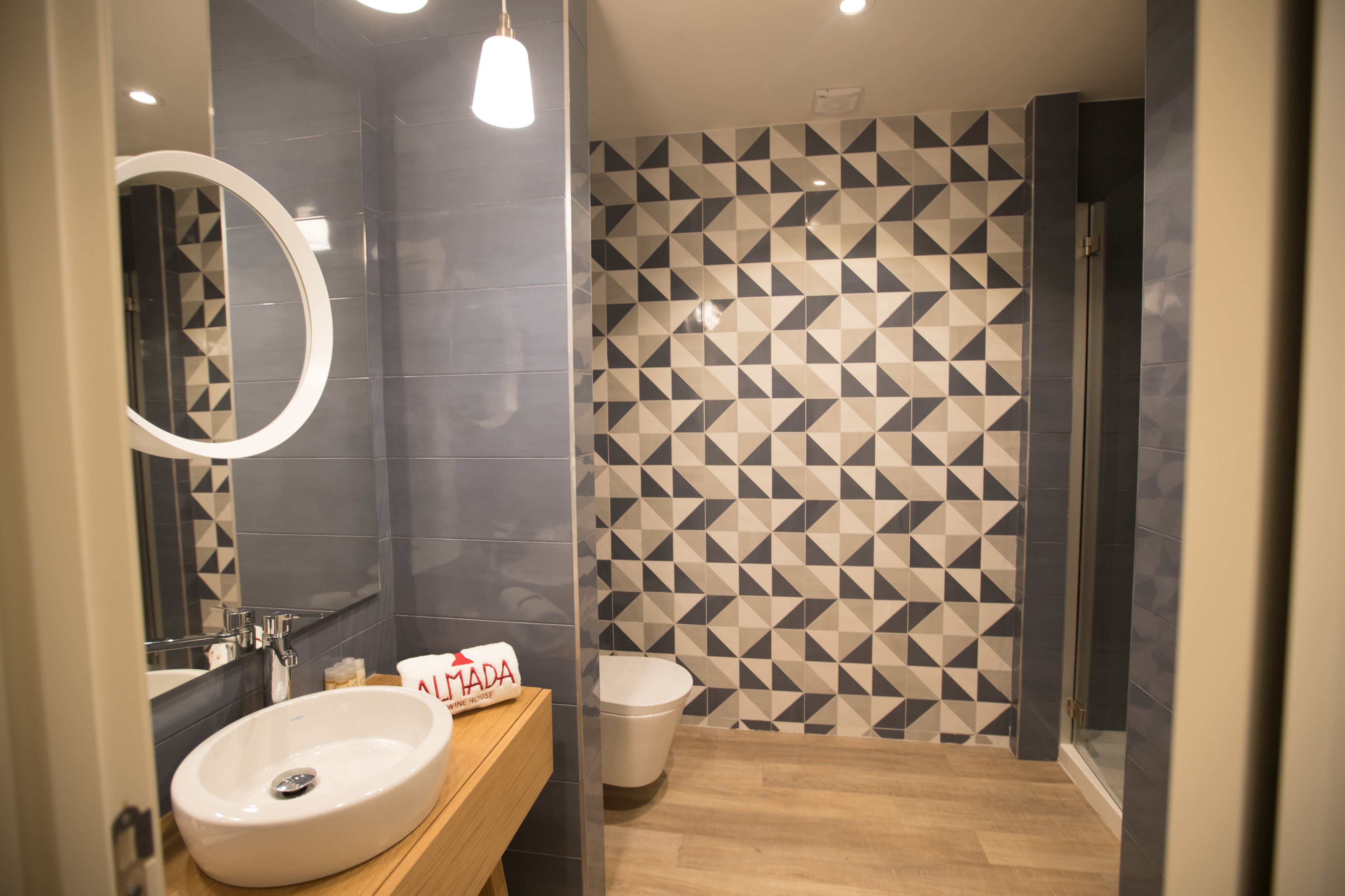 A modern bathroom with a round mirror, a sink, and a toilet. The walls have a geometric tile pattern.