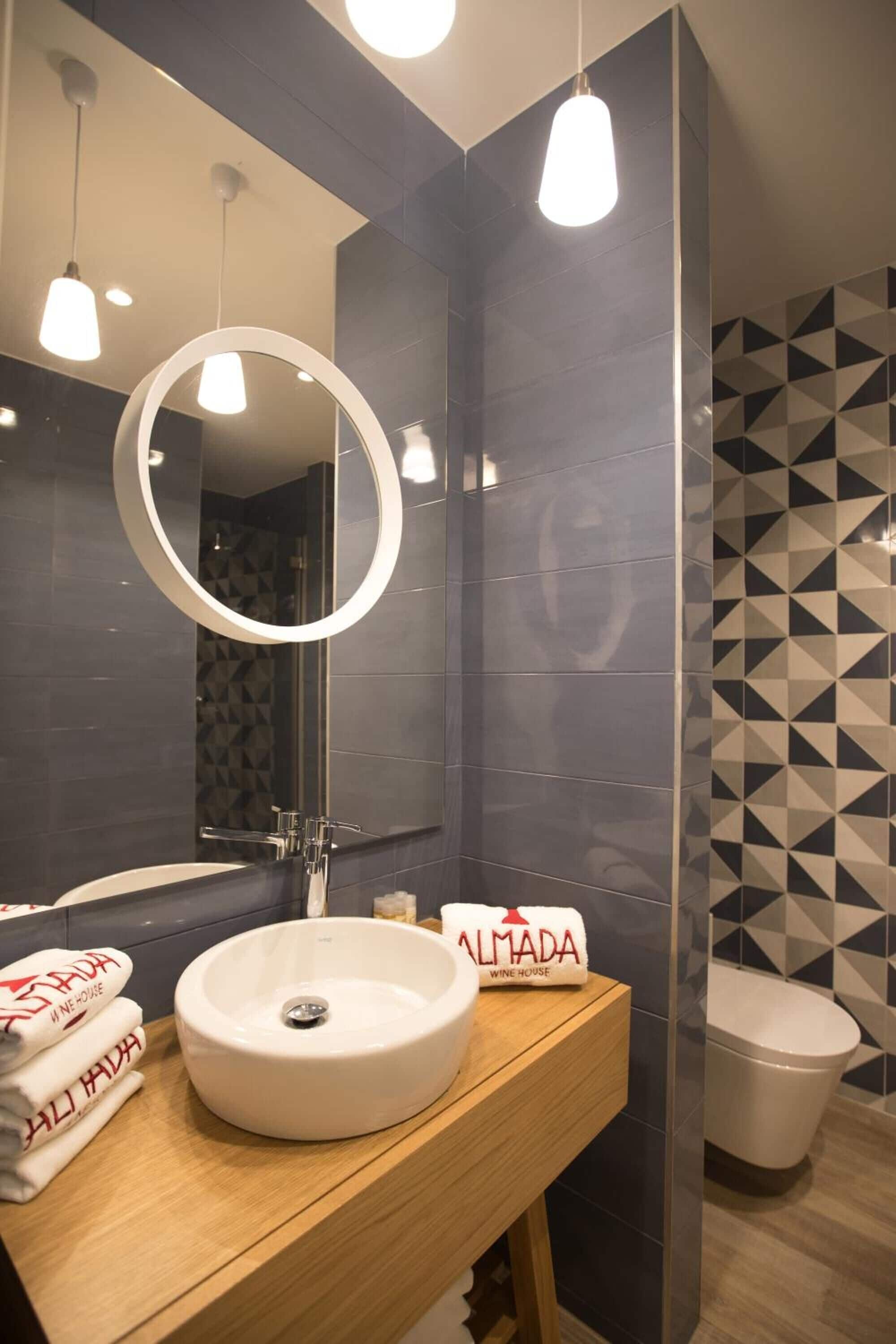A modern bathroom with a round mirror, a sink, and a toilet. There are towels on the sink and a sign that reads "ALMADA WINE HOUSE".