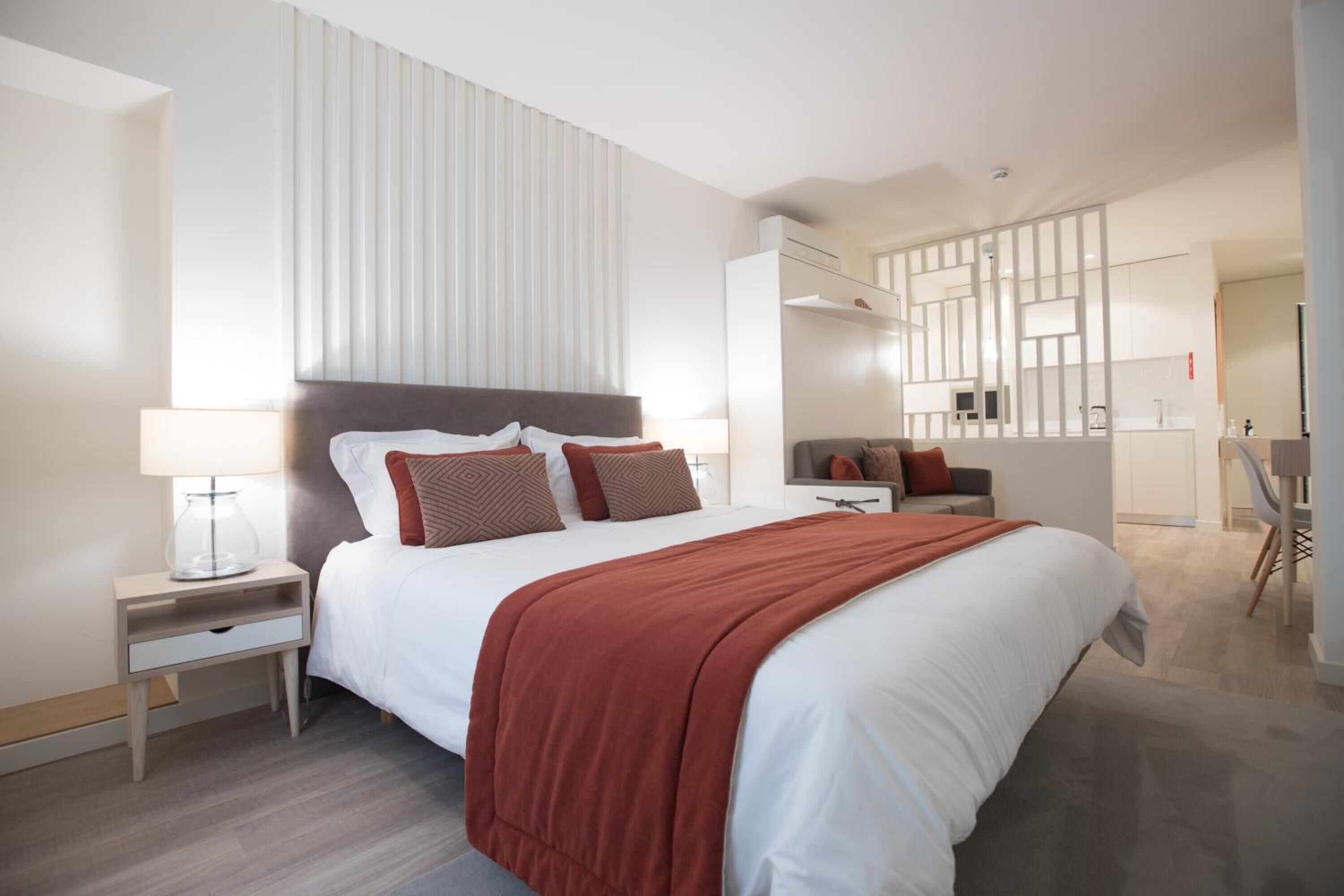 A modern hotel room with a large bed, a bedside table with a lamp, a small desk, and a chair.
