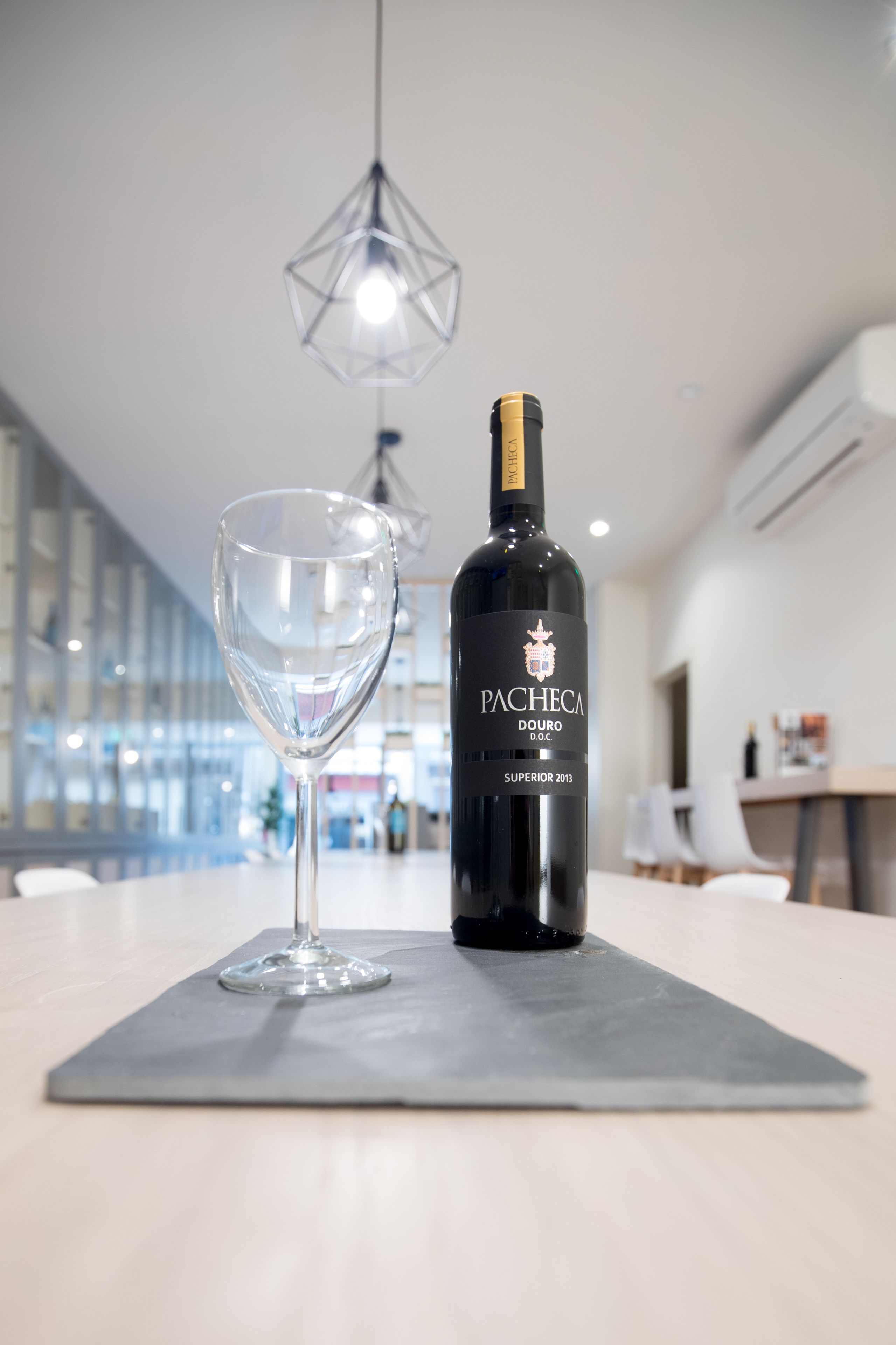 A bottle of Pacheca Douro D.O.C. wine and a wine glass on a table in a modern interior with a geometric light fixture.