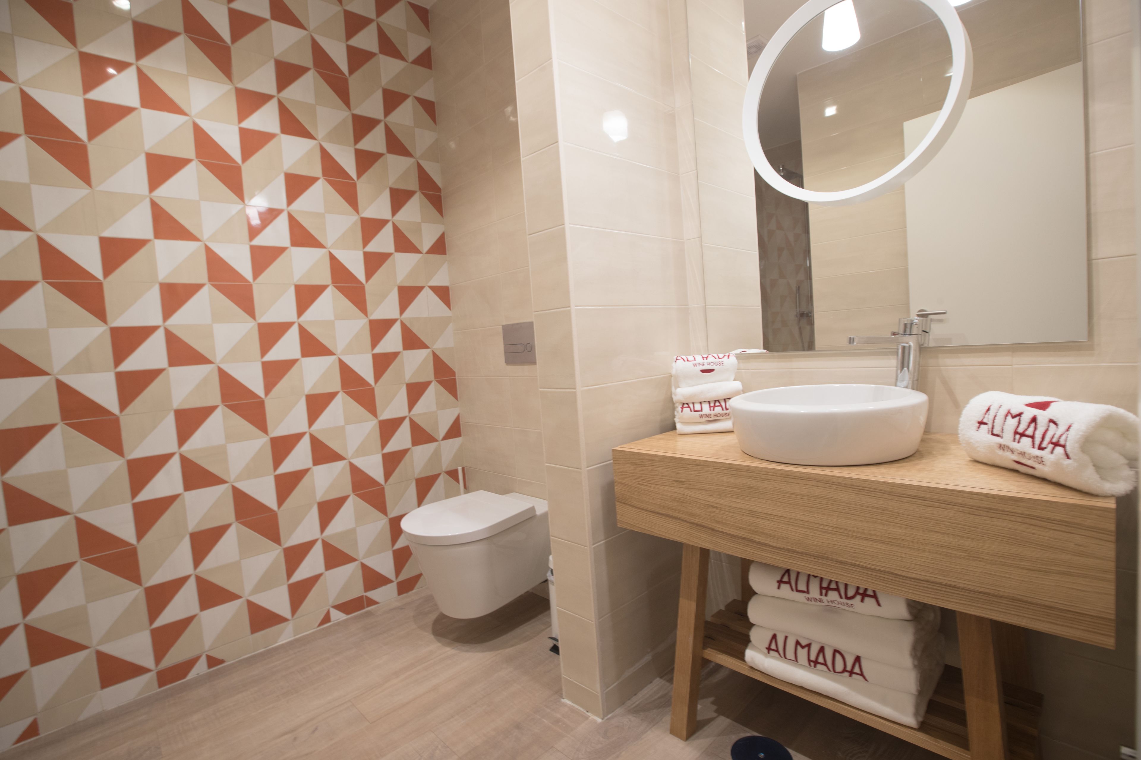 Une salle de bain avec un mur orné d’un motif géométrique, des toilettes, un lavabo avec un miroir et des serviettes de la marque « Almada ».