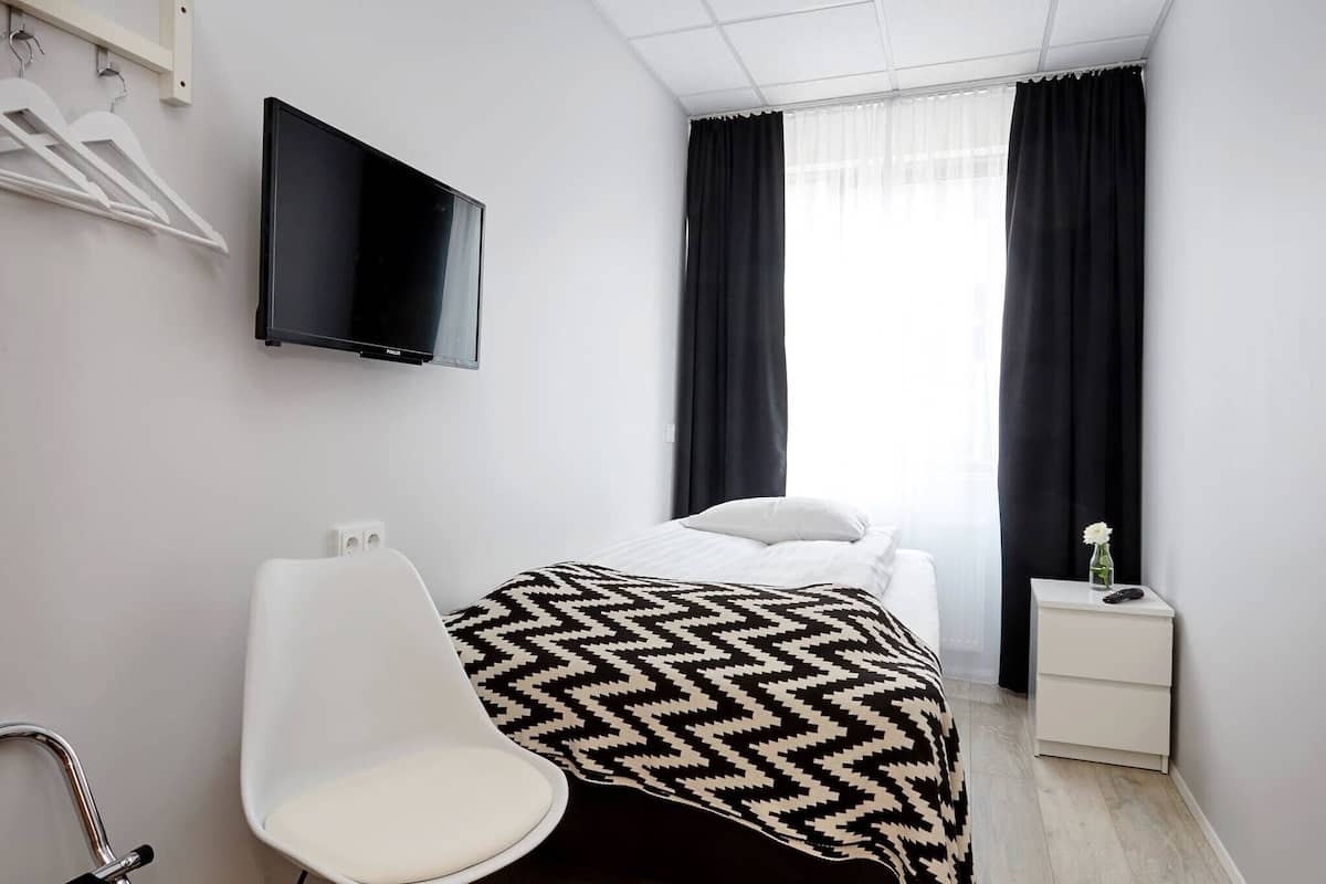 A hotel room with a bed, a TV mounted on the wall, a chair, a bedside table with a vase, and a window with curtains.