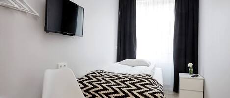 Ein Hotelzimmer mit einem Bett, einem an der Wand befestigten Fernseher, einem Sessel, einem Nachttisch mit einer Vase und einem Fenster mit Vorhängen.